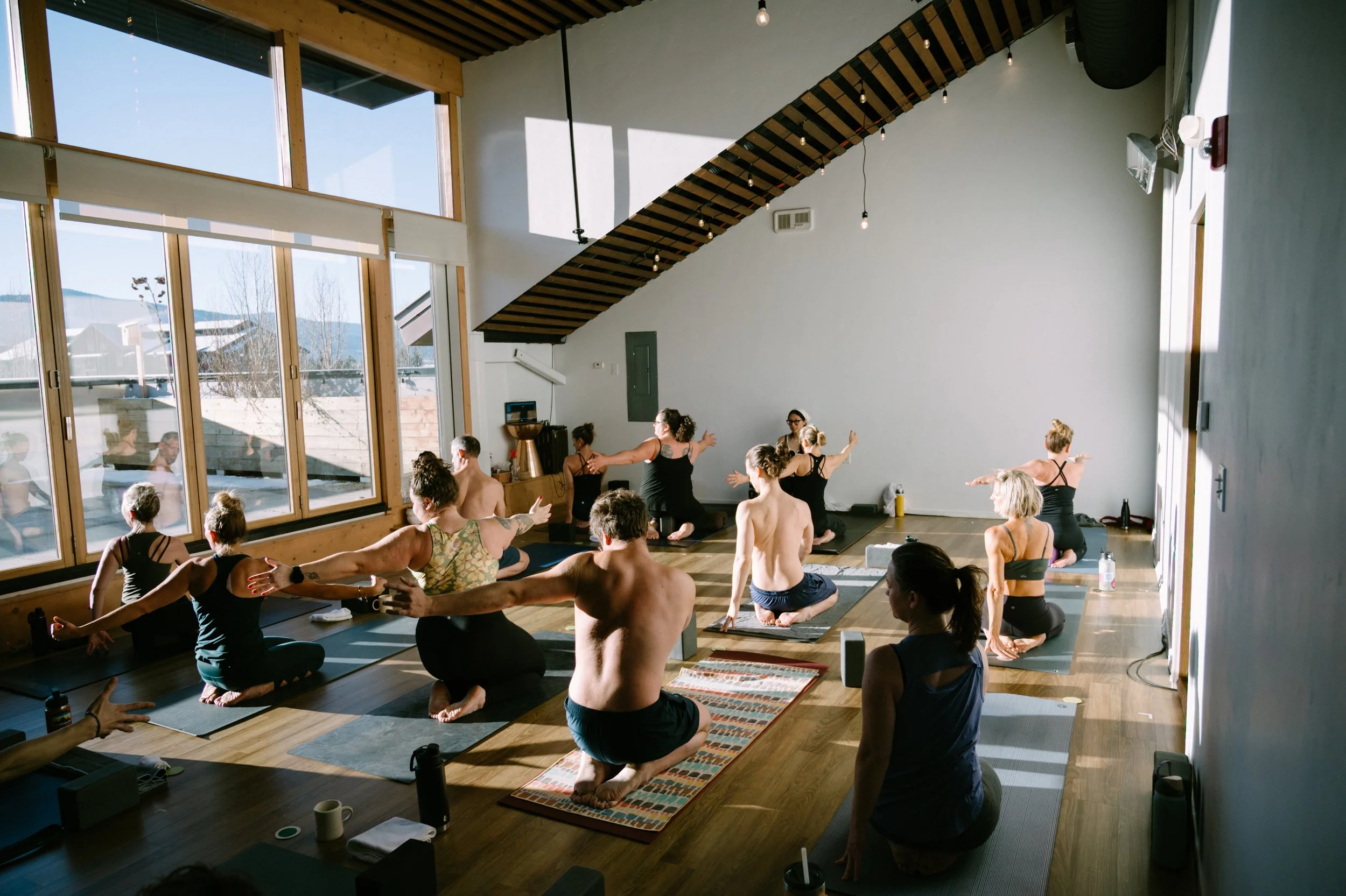 A group of people practicing yoga in a bright studio with large windows and wooden floors.