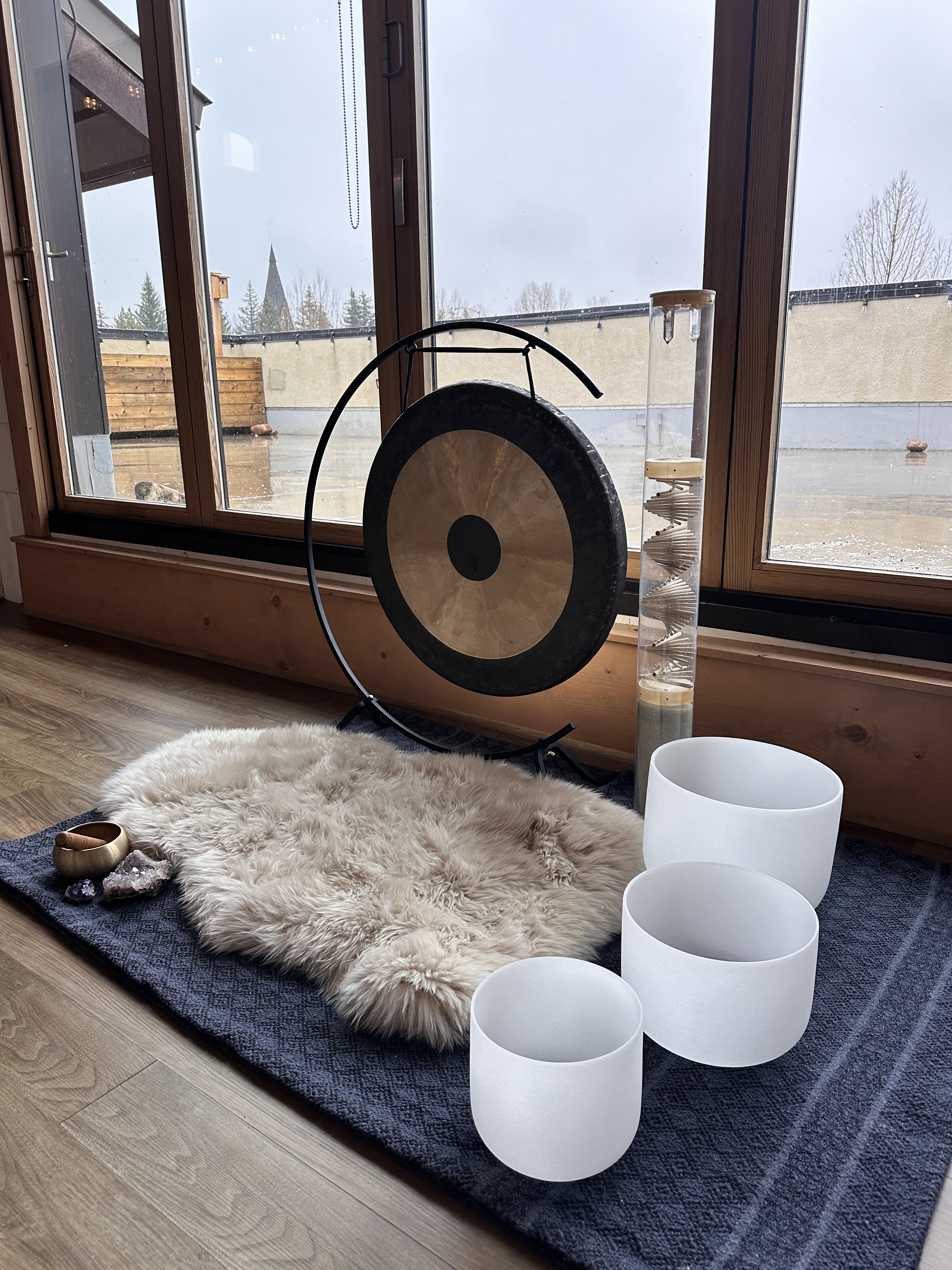 Interior view of a room near a large window showing an outdoor patio and overcast sky. Inside, there is a fluffy white rug with several crystals and a singing bowl, white crystal singing bowls, a vertical water fountain, a shell, and a fluffy throw blanket. The room features wooden trim and flooring.