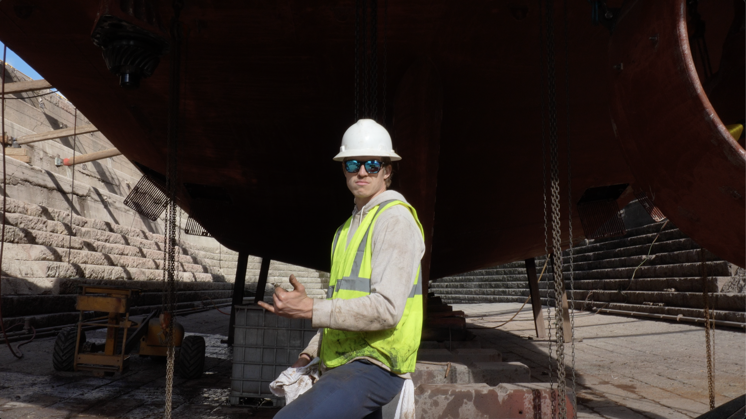 A construction worker wearing a white safety helmet, reflective vest, and sunglasses standing under a ship's hull at a shipyard.