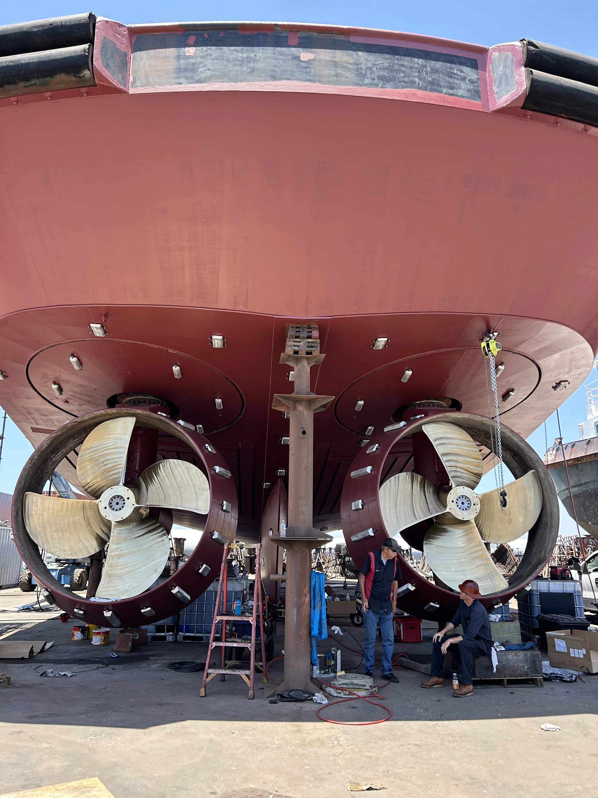 Close-up view of a large ship's hull with two large wooden propellers underneath. The ship is on dry dock, with workers and equipment around working on the vessel.