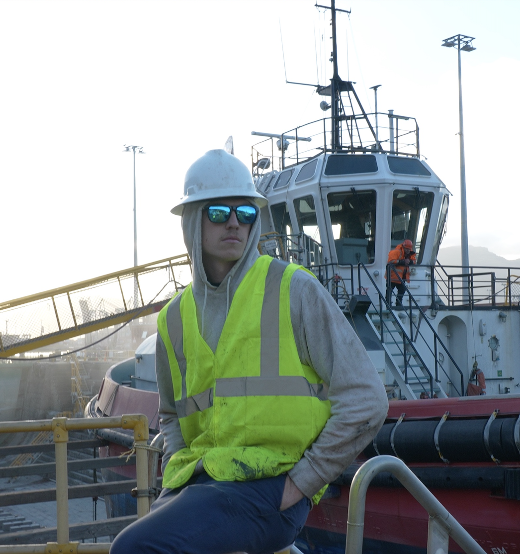 A construction worker with sunglasses, a white hard hat, and a yellow high-visibility vest stands on a dockside near a ship, with another worker in orange clothing visible on the ship in the background.