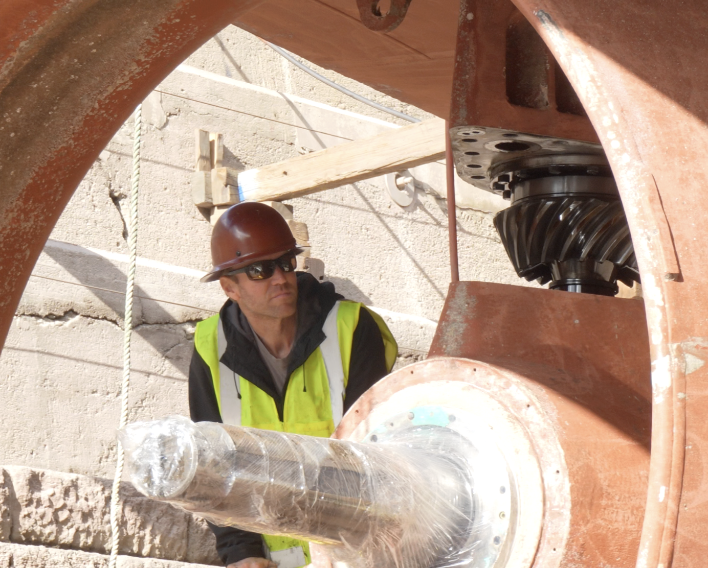 A construction worker wearing a brown hard hat, sunglasses, and a yellow safety vest working near a large industrial gear inside a construction site with concrete walls and wooden supports.
