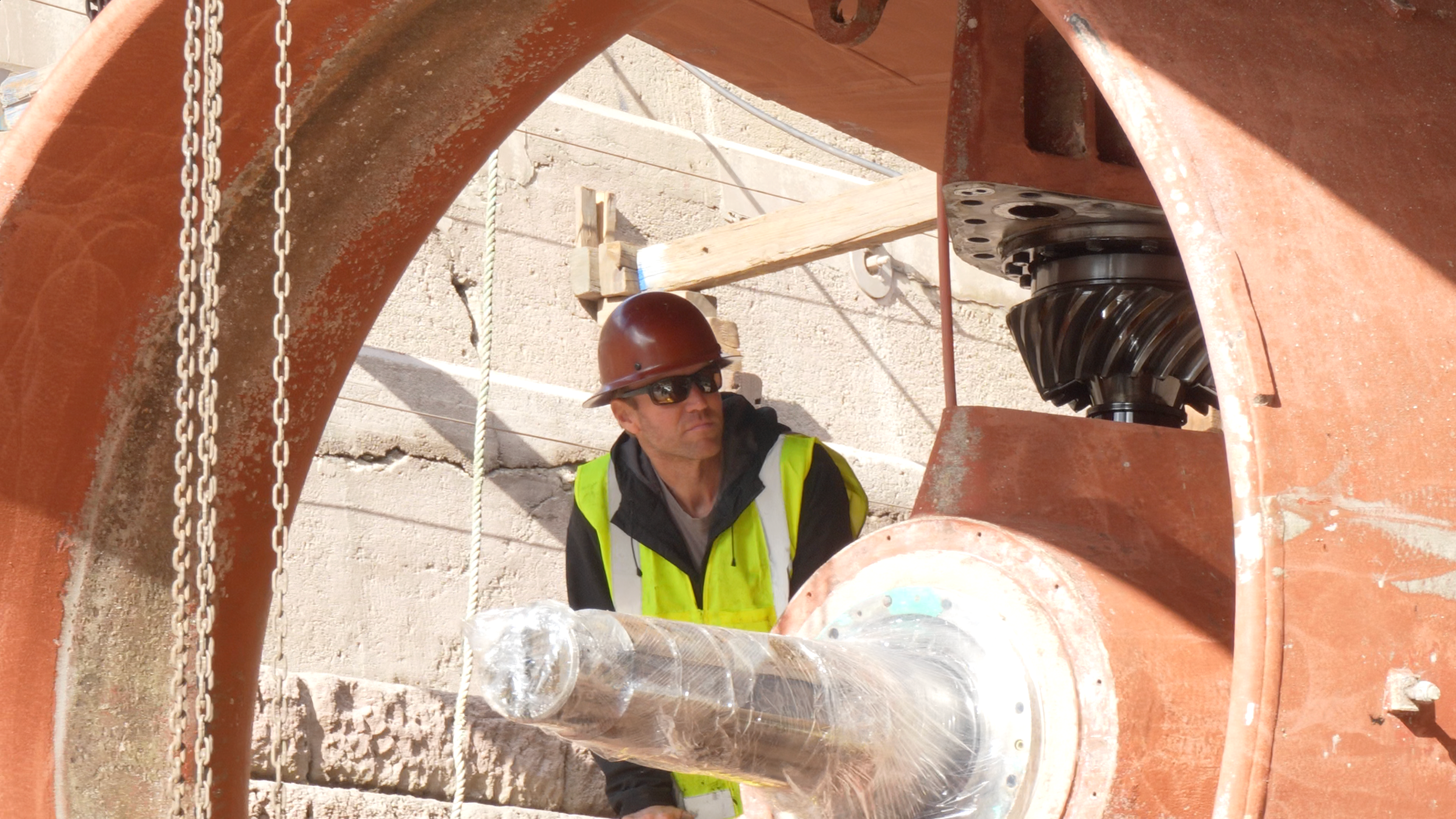 A construction worker wearing a brown hard hat, sunglasses, a yellow safety vest, and a black hoodie working on large machinery at a construction site, with concrete wall in the background.