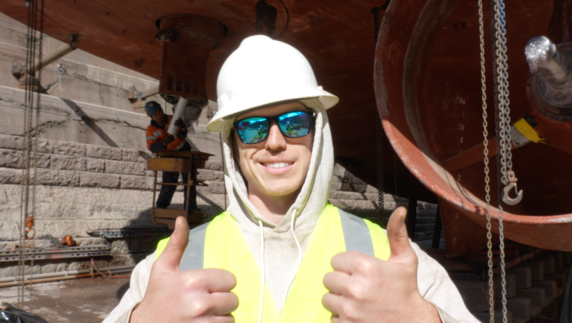 Close-up of a smiling construction worker wearing a white hard hat, sunglasses, and a yellow safety vest, giving two thumbs up at a construction site with a large red boat hull in the background.