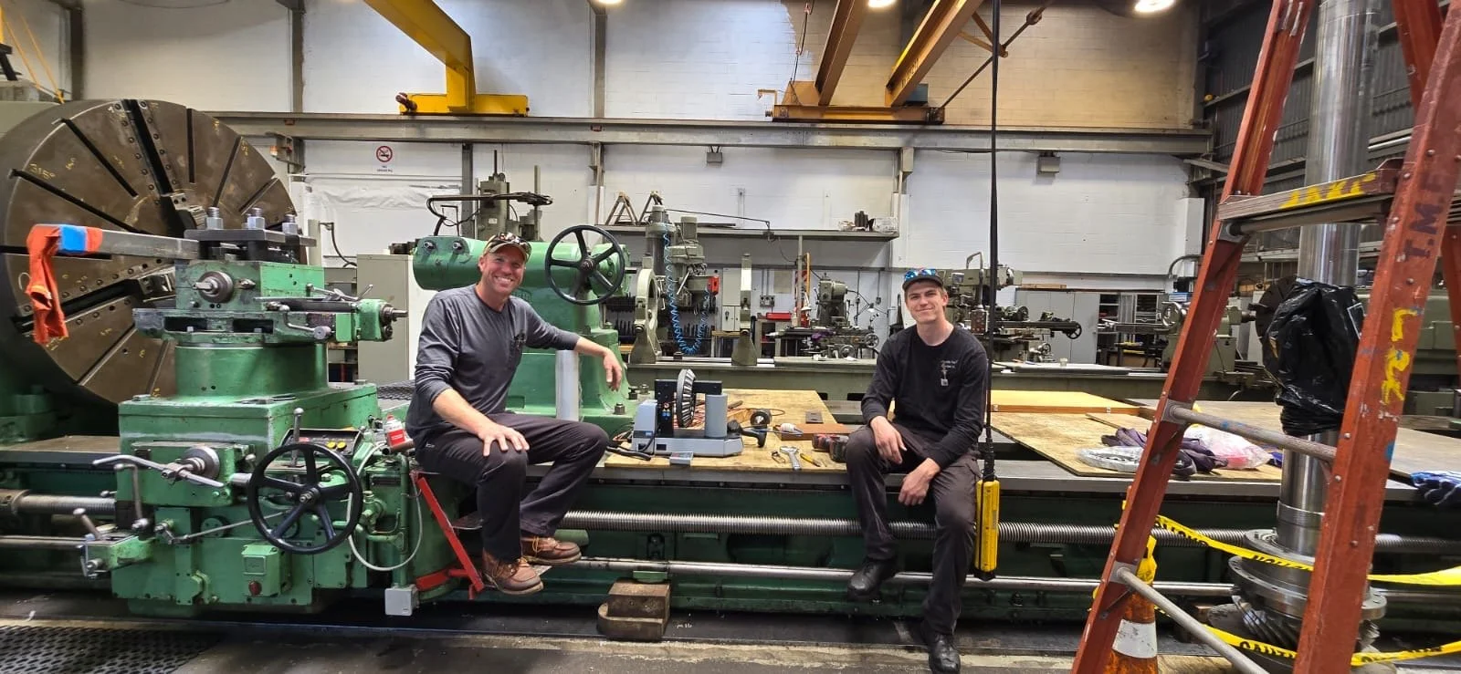 Two men sitting on a large green industrial machine inside a factory or workshop. The machine has various dials and parts, with tools and equipment on a wooden table between the men. In the background, there are more machines and industrial equipment, with a ladder on the right side and caution tape.