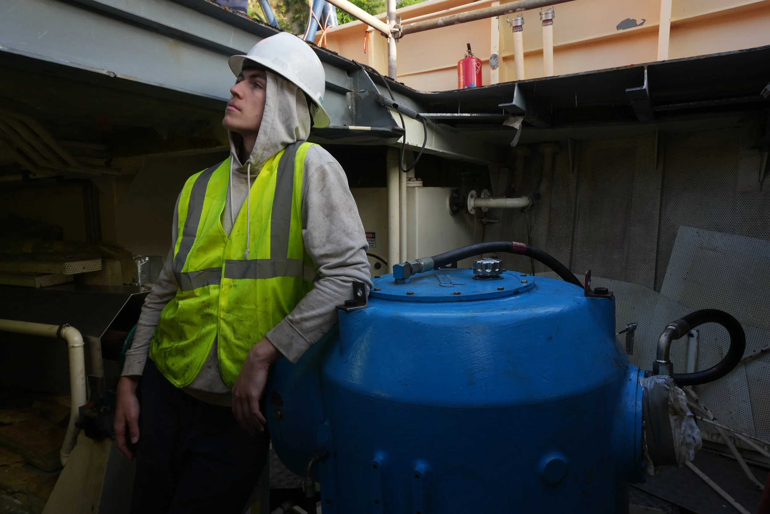A man wearing a white safety helmet, a gray hoodie, and a yellow safety vest leans against a large blue industrial tank at a construction or industrial site, with pipes and scaffolding visible in the background.