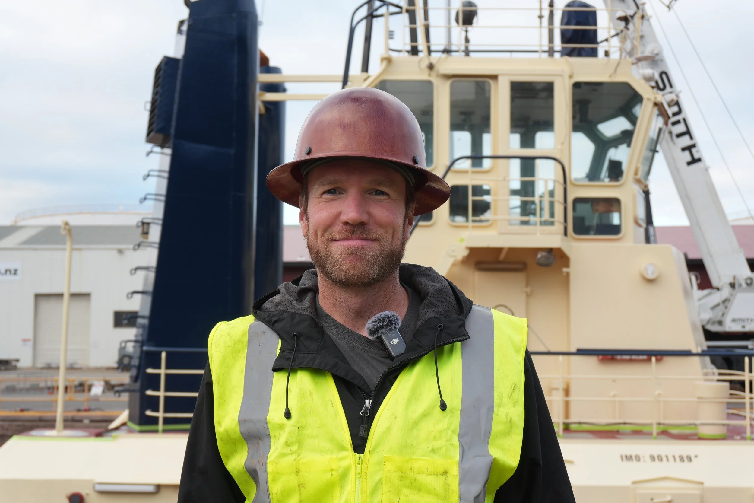 A construction worker wearing a safety helmet, sunglasses, and a high-visibility vest working near a large mechanical gear or propeller shaft.