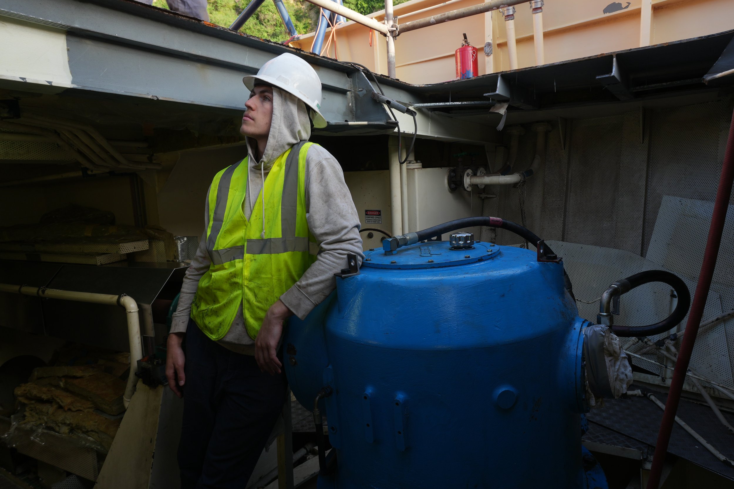 A man wearing a white safety helmet, gray hoodie, and yellow safety vest stands next to a large blue industrial tank inside a mechanical room with pipes and equipment.