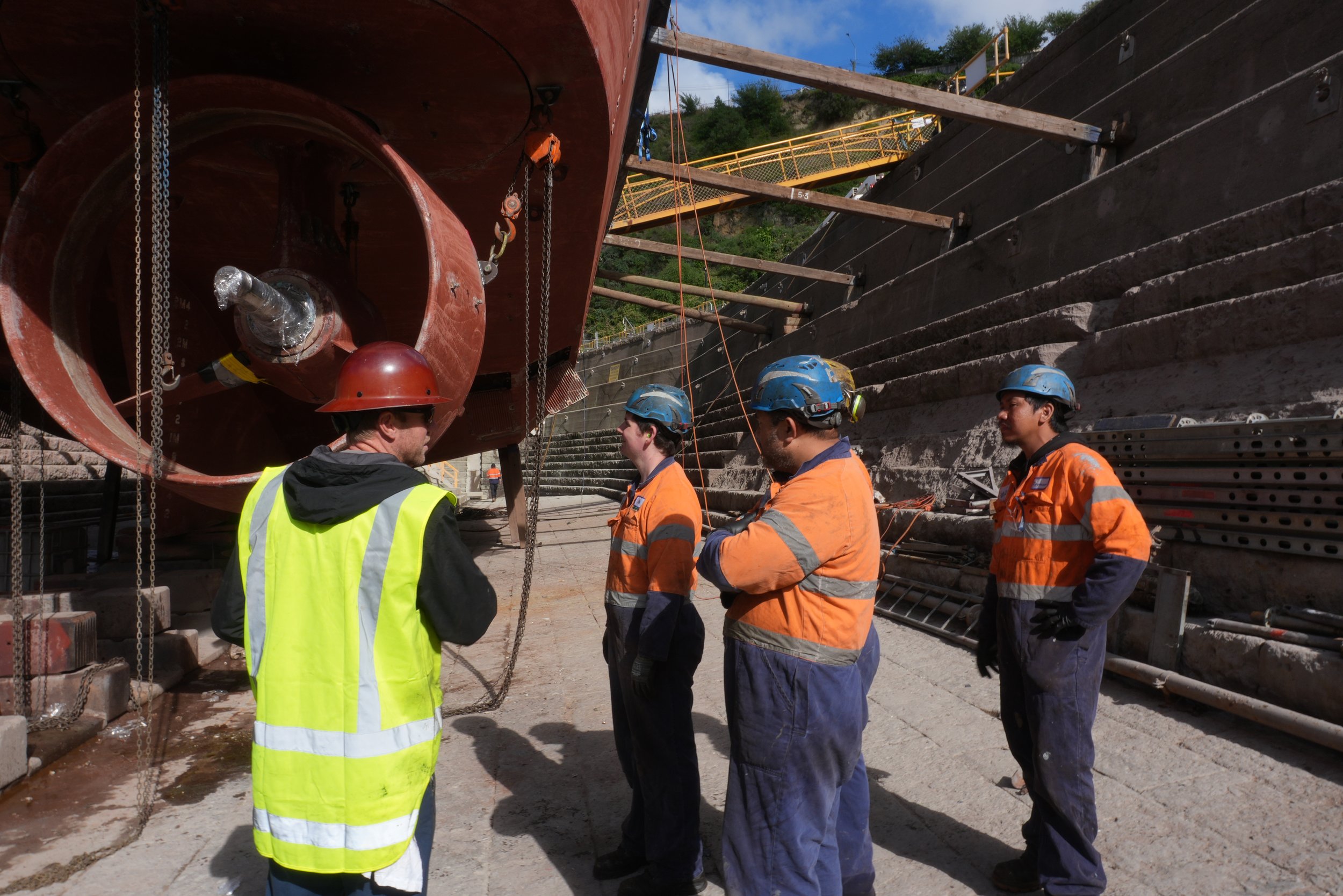Construction workers, including a person in a red helmet and yellow safety vest, stand near a large, partially submerged pipe on a construction site, with a concrete wall and hillside in the background.