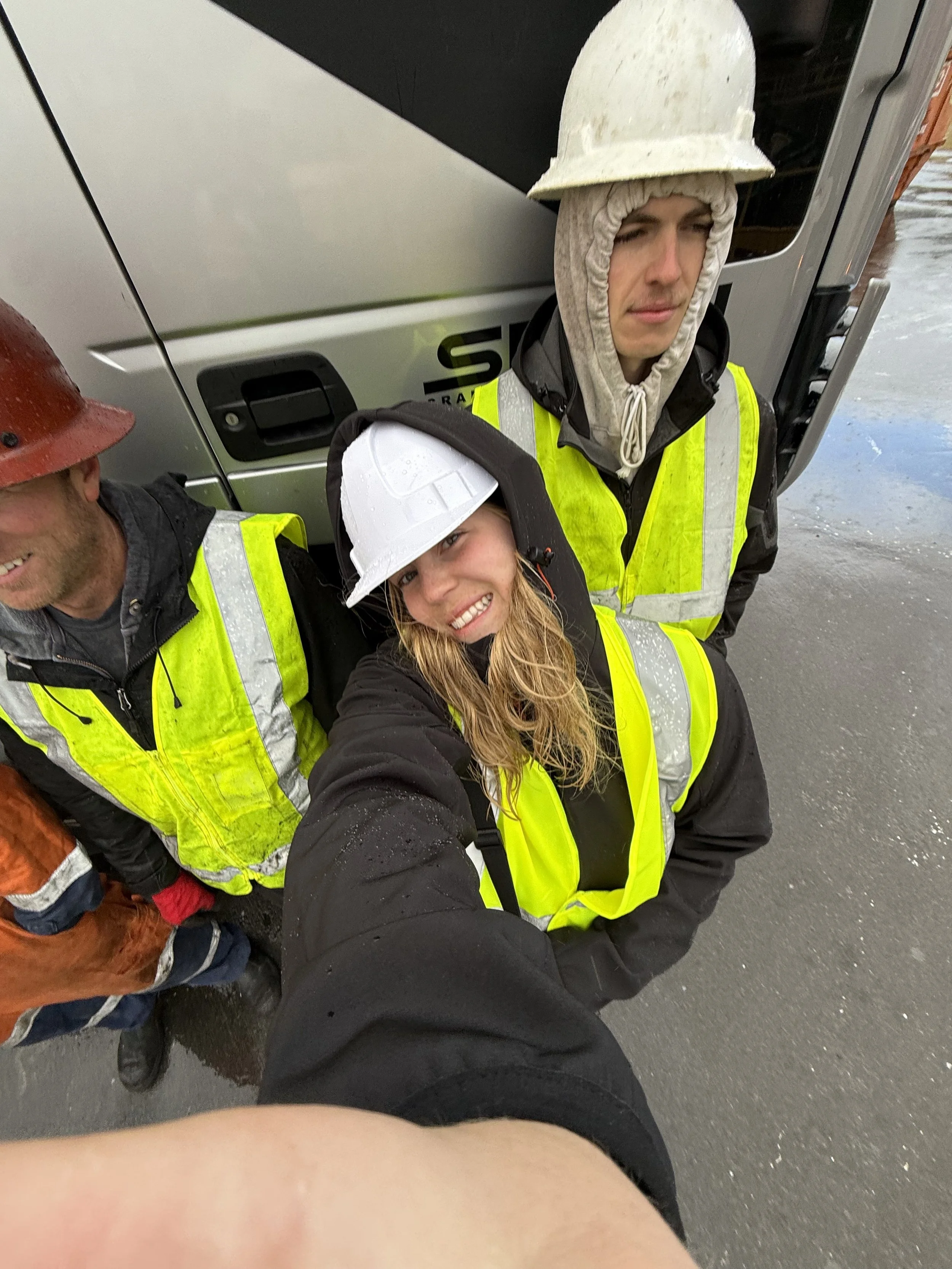 Group of construction workers taking a selfie outdoors in wet weather, wearing high-visibility safety vests and hard hats, with a truck in the background.