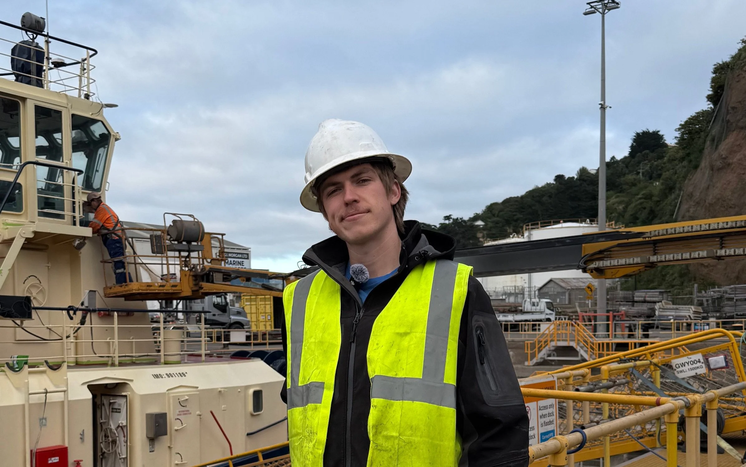 A construction worker with sunglasses, a hard hat, a hoodie, and a yellow safety vest giving a thumbs up at a boat repair site. Two other workers are visible in the background working on the hull of a boat that is supported by wooden blocks.