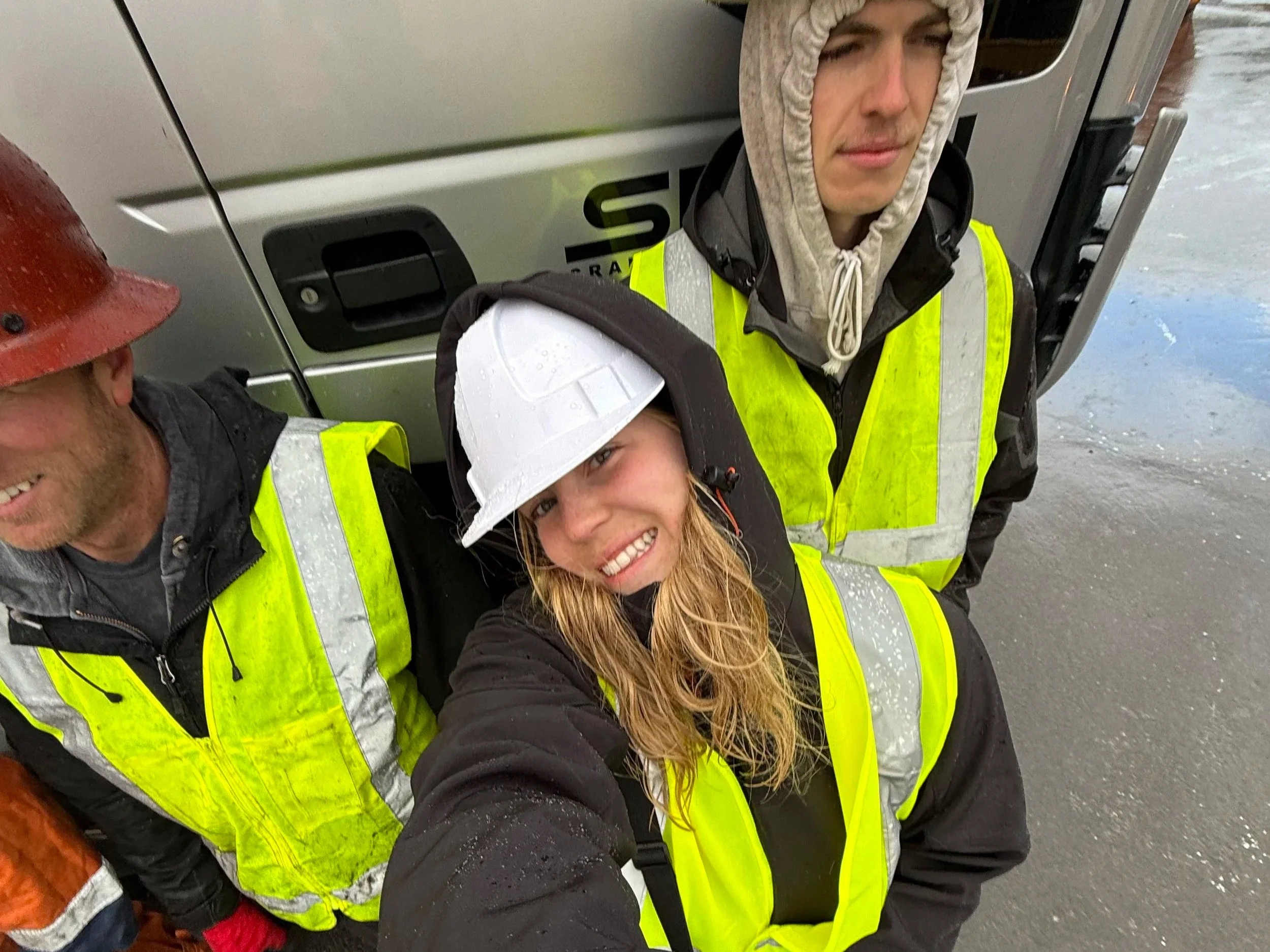 Three construction workers wearing high-visibility vests and hard hats, standing outdoors near a large truck on a wet surface.