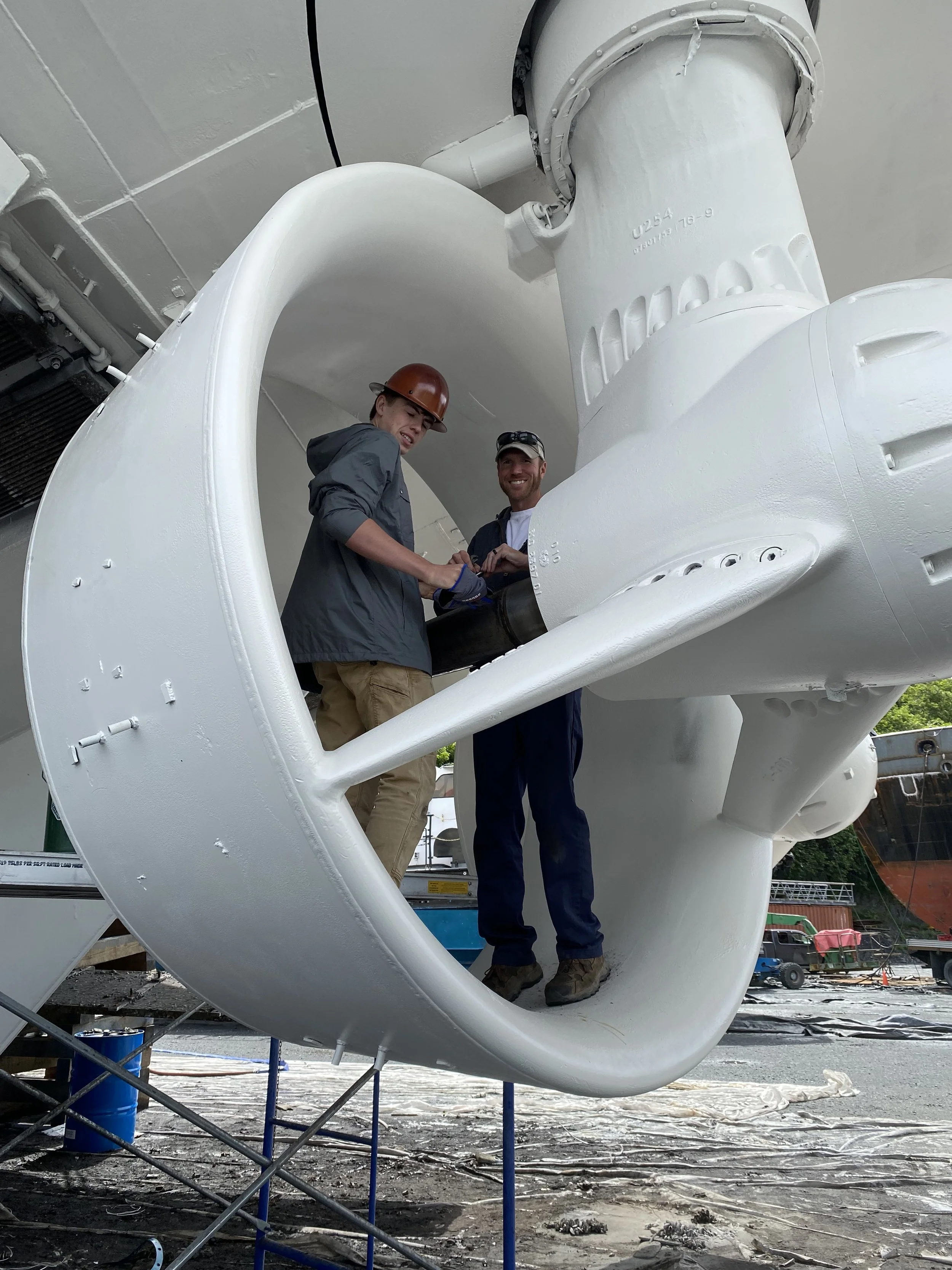 Two workers inside a large ship's propeller, with one wearing a safety helmet, working on or inspecting the machinery.