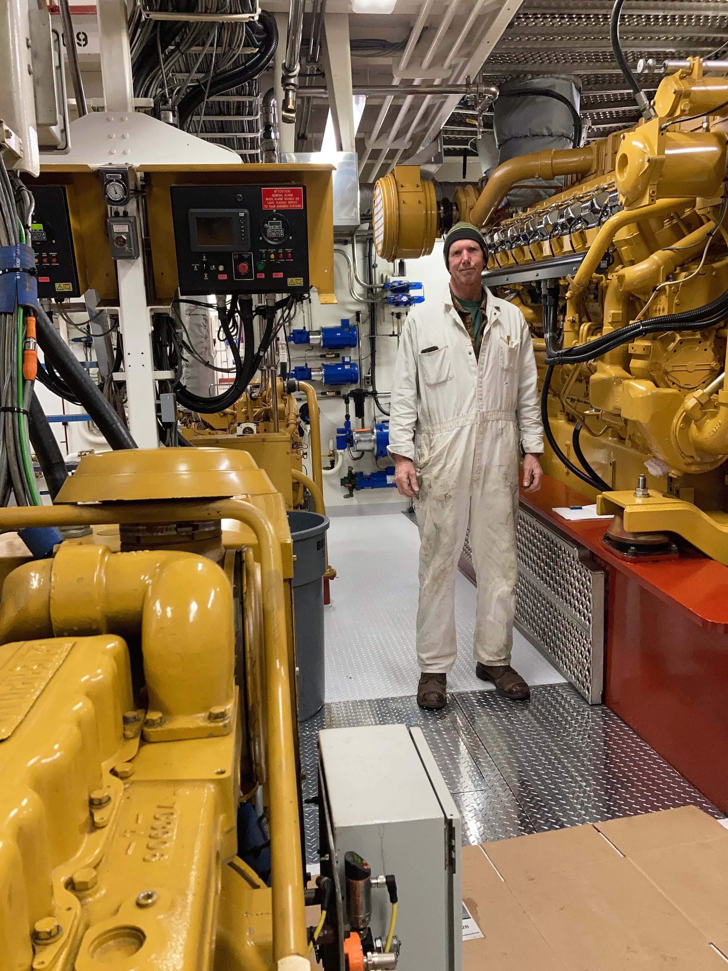 A middle-aged Asian man with gray hair sitting on a beige bench against a light-colored wall, wearing a blue short-sleeved sweater and beige pants, looking to his left with a calm expression.