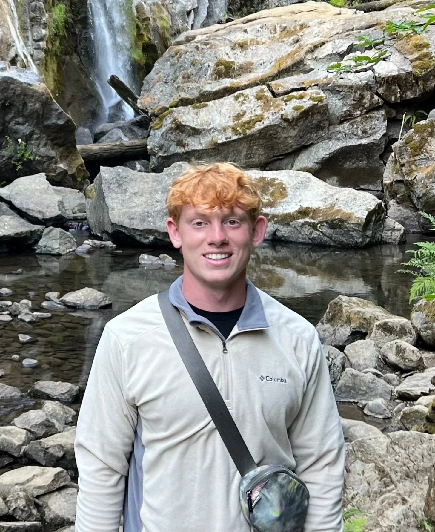 A red-haired young man smiling in a rocky outdoor setting with a small waterfall and stream in the background.