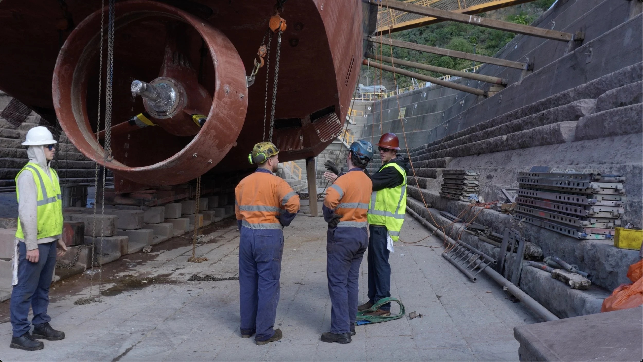 Construction workers in safety gear inspecting a large boat hull at a shipyard, with scaffolding and construction materials around.