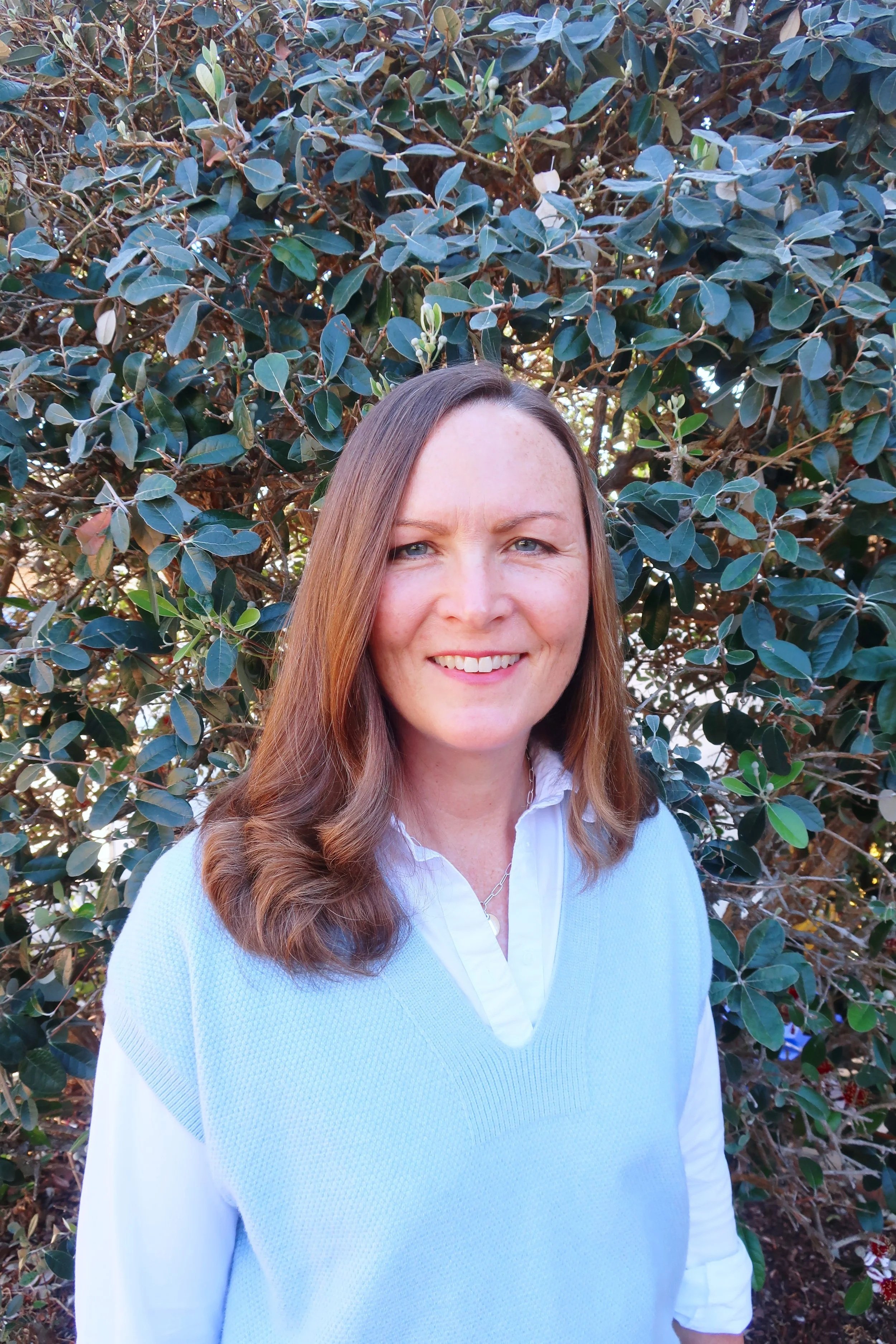 A woman with long brown hair, wearing a white shirt and a light blue vest, standing outside in front of green shrubbery.