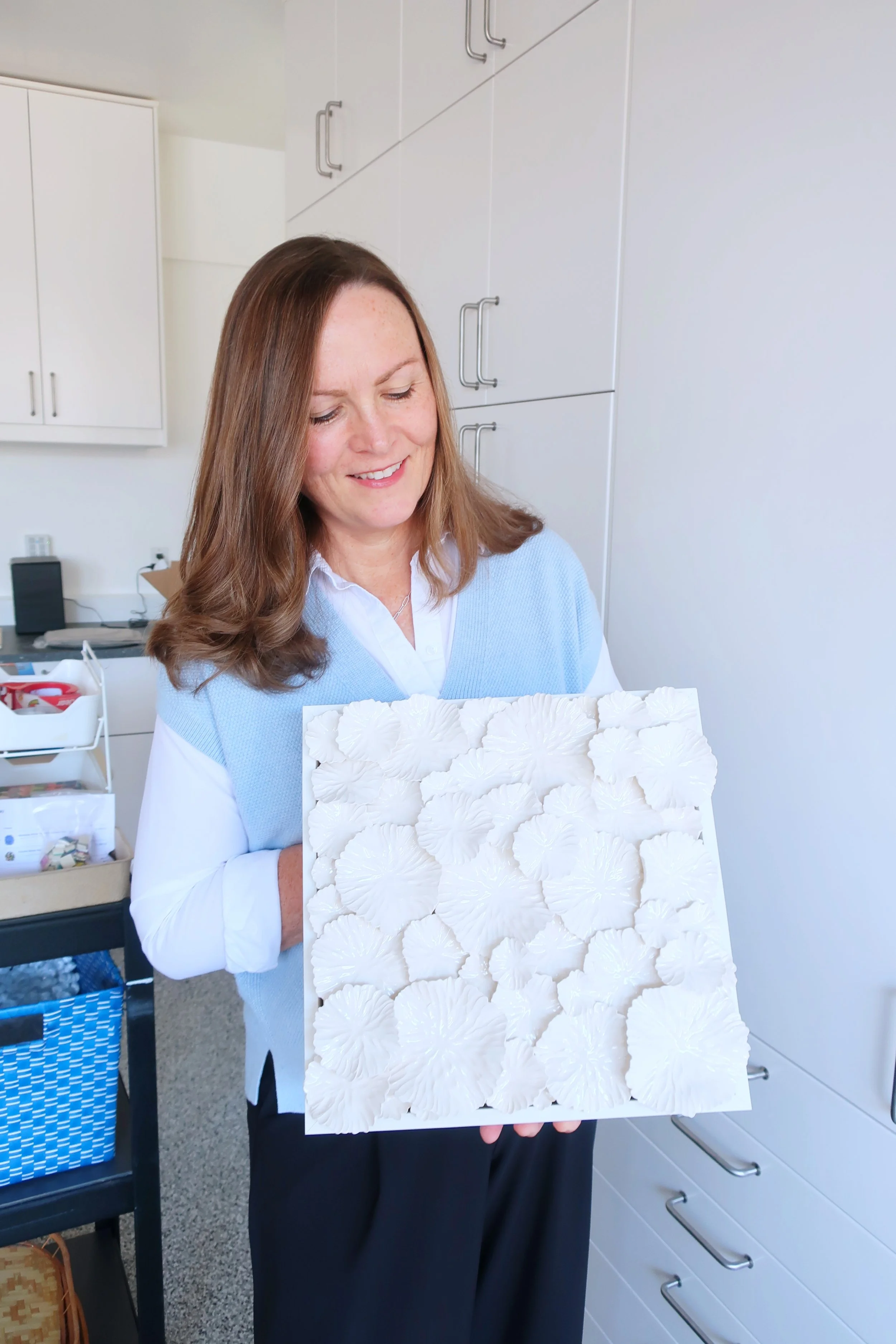 A woman with brown hair smiling and holding a white textured art piece with a floral pattern indoors, with white cabinets and a cart in the background.