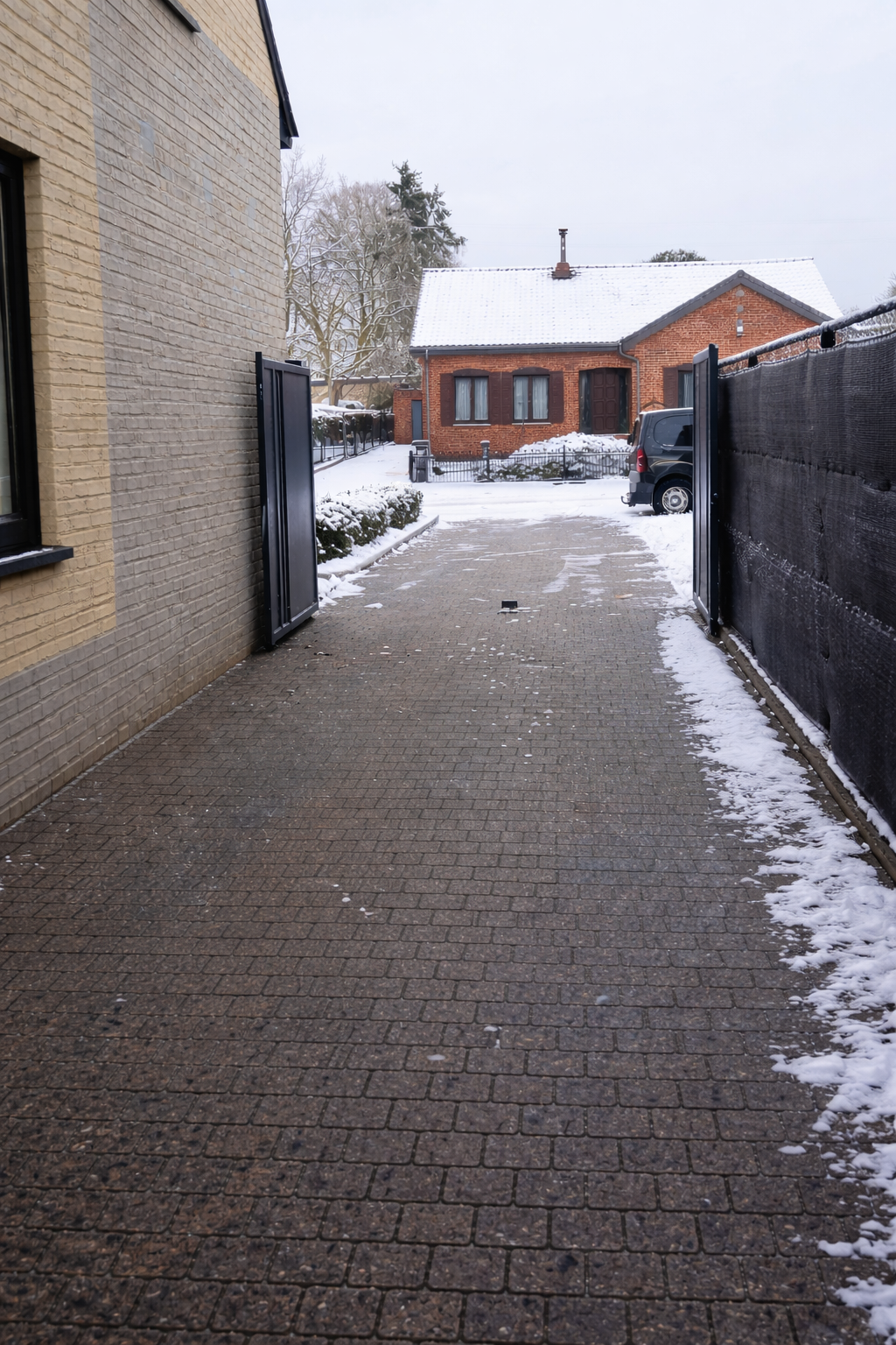 Een oprit met een betegeld pad dat bedekt is met een dun laagje sneeuw. Aan beide zijden van de oprit staan hekken; links een open metalen poort, rechts een zwarte hek. Op de achtergrond staat een rode bakstenen huis met een sneeuwbedekt dak en een zwart getint raampartij.