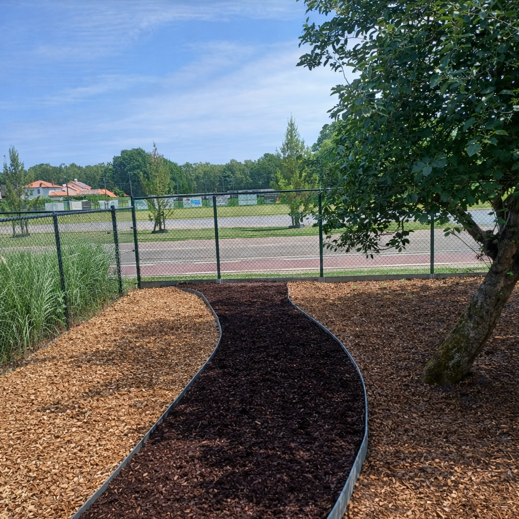 Tuin met een bochtige pad, omringd door een boom en een hek, met grasvelden en huizen op de achtergrond onder een blauwe lucht.