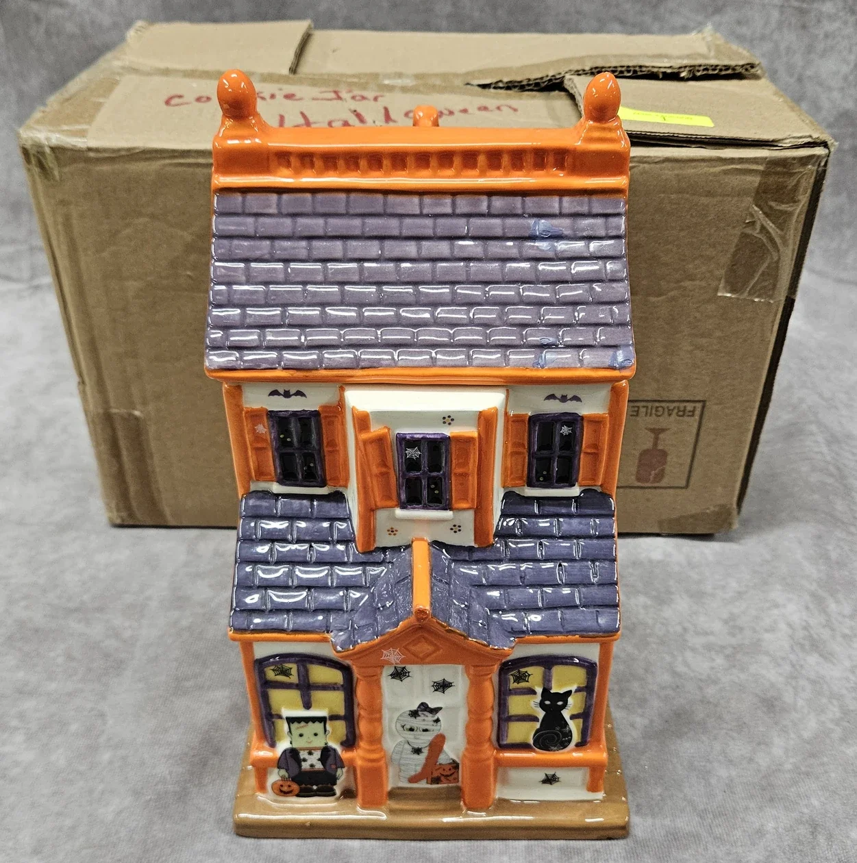 Decorative ceramic Halloween house figurine with orange and purple accents, windows, a front door, and Halloween decorations, and a grey tiled roof.