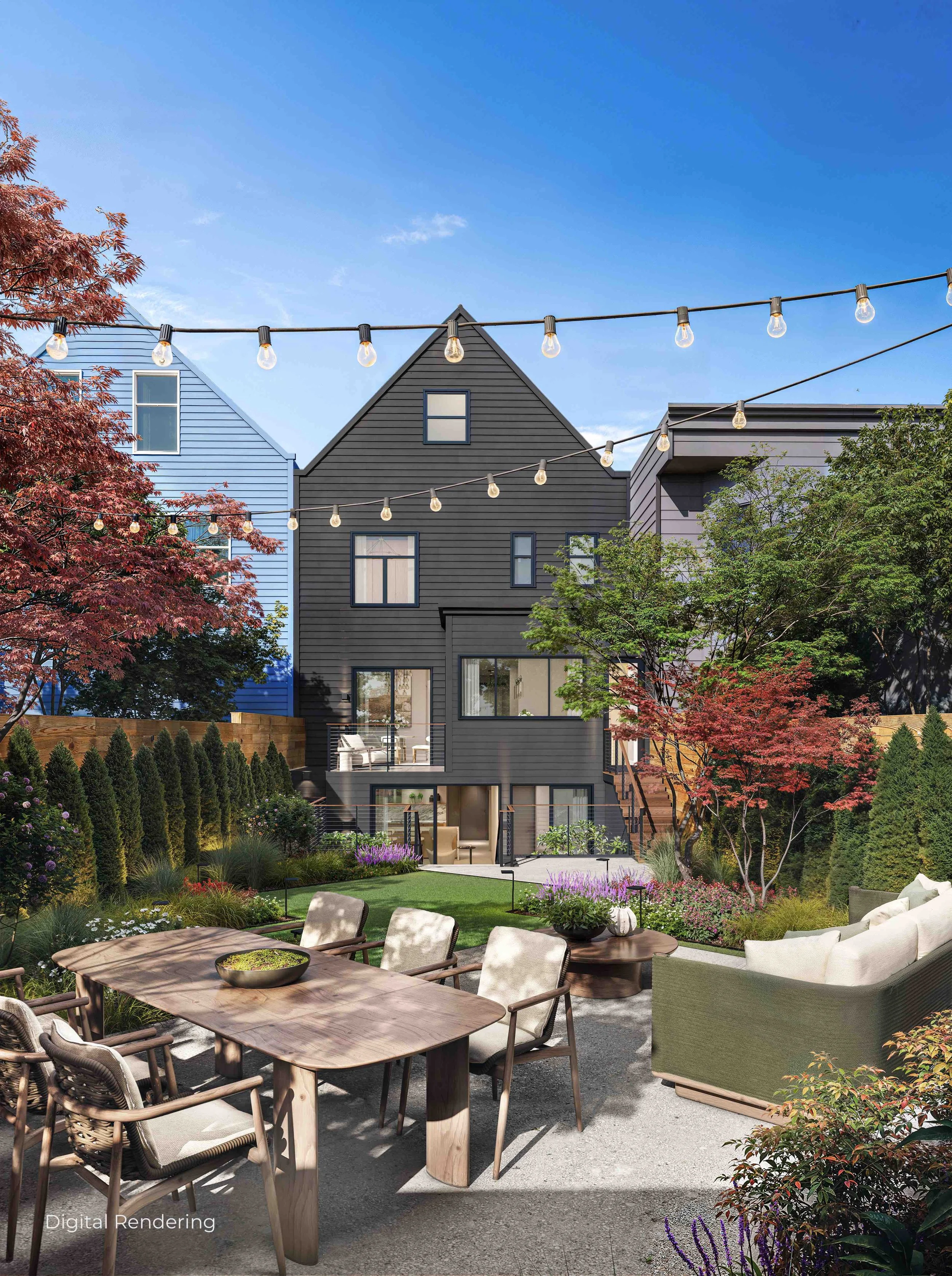 An outdoor garden patio area with a wooden dining table, chairs, and lush greenery, including trees with red and green leaves. String lights are hanging overhead, and modern houses are visible in the background under a clear blue sky.