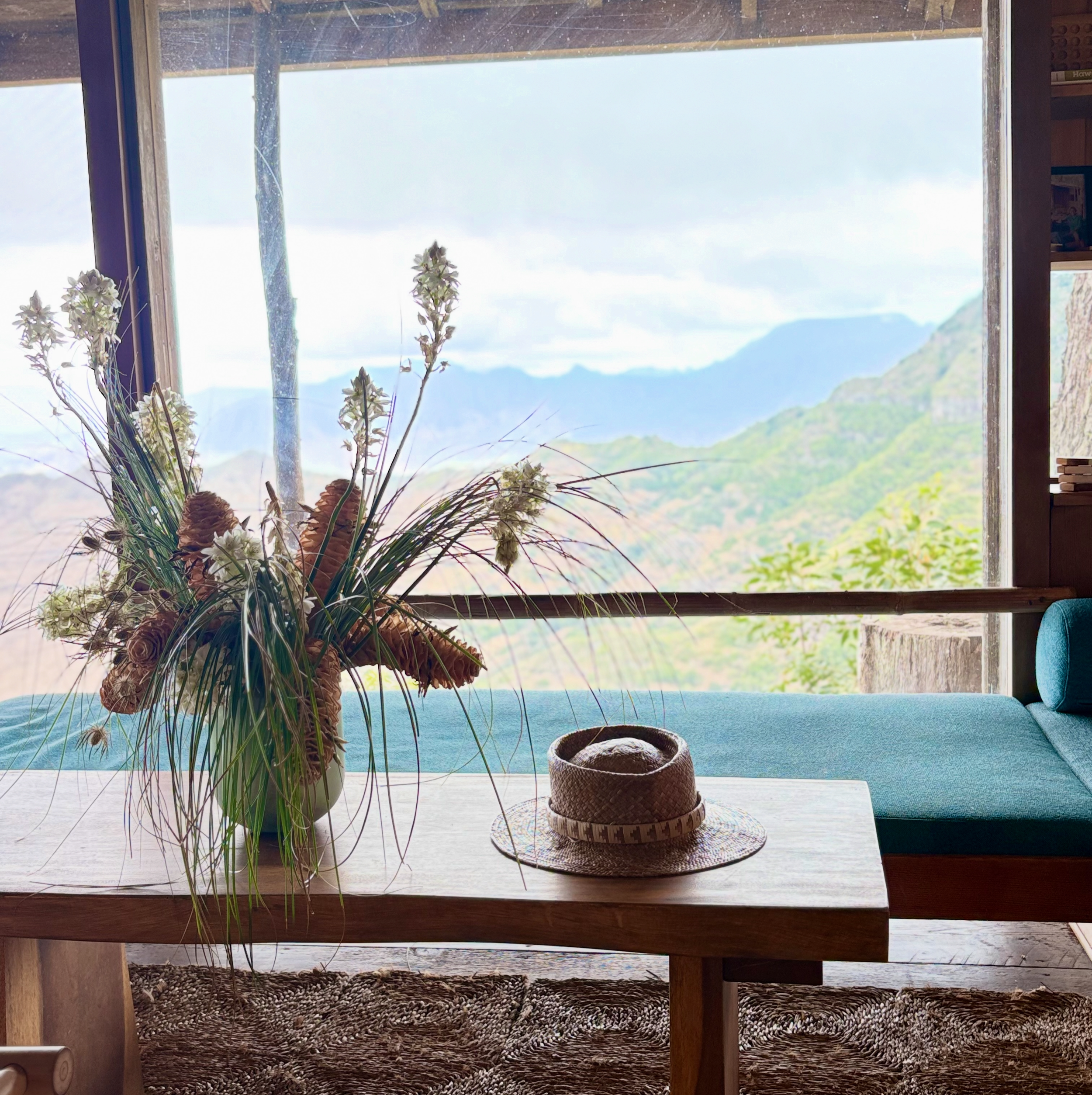 A wooden table with a large flower arrangement in a vase and a straw hat. A window in the background shows mountains and a cloudy sky.