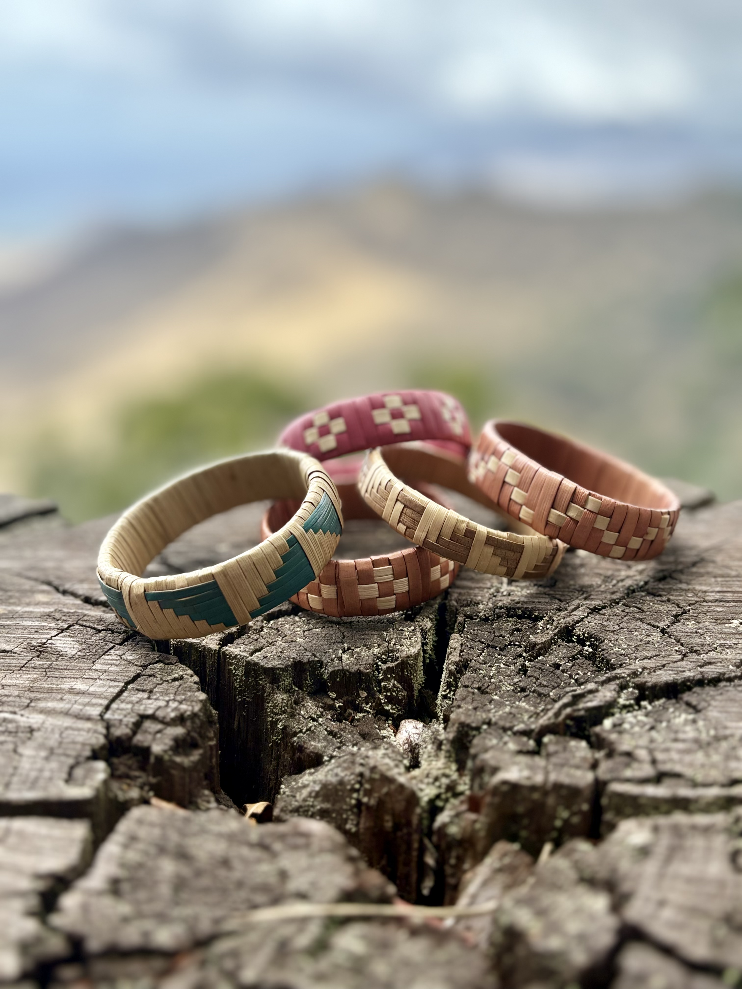 Four woven bracelets in different colors are placed on a weathered wooden log outdoors with blurred landscape background.