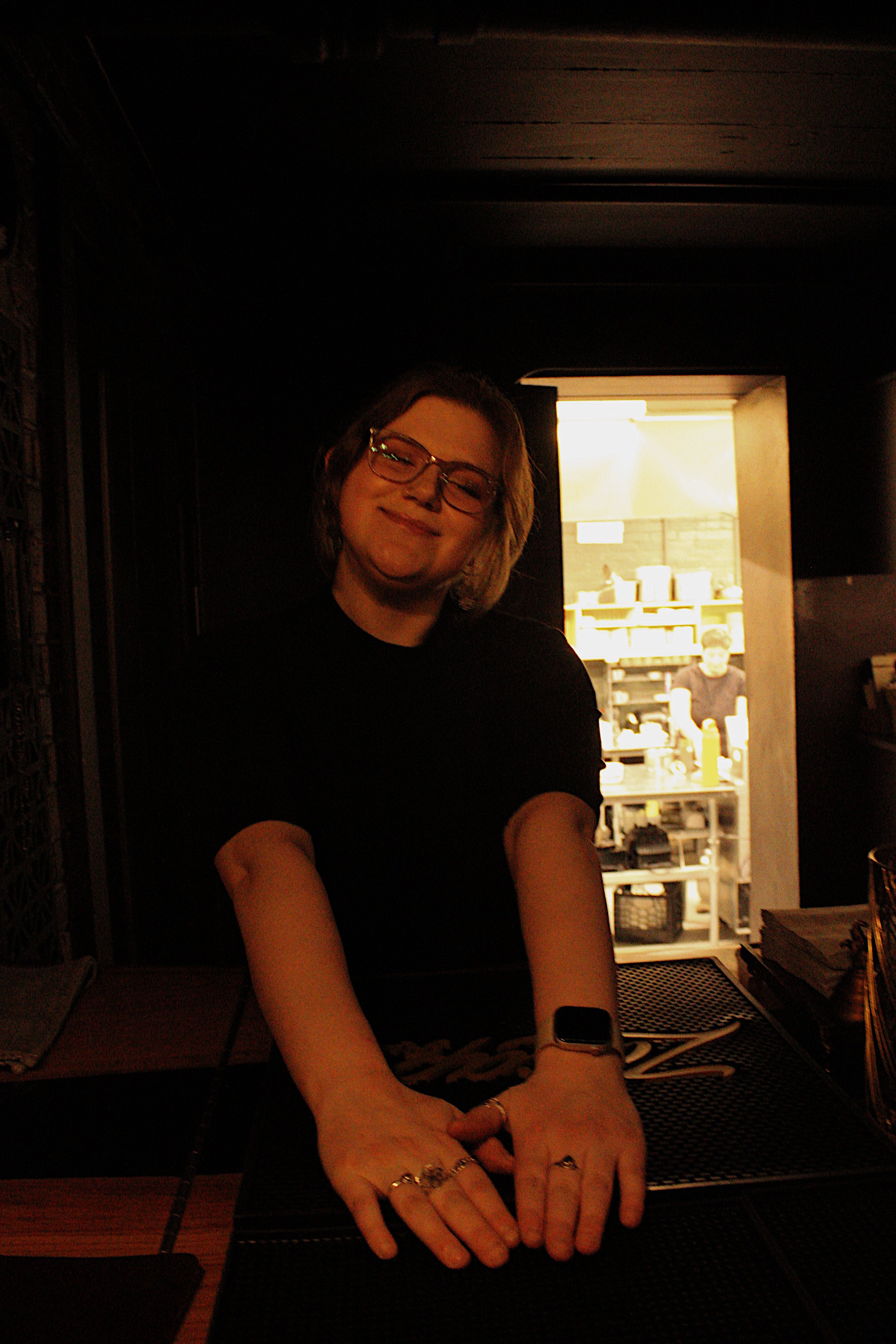 A woman with glasses, blonde hair, and wearing jewelry is smiling and leaning on a table in a dimly lit room, with a brightly lit kitchen or workspace visible in the background.