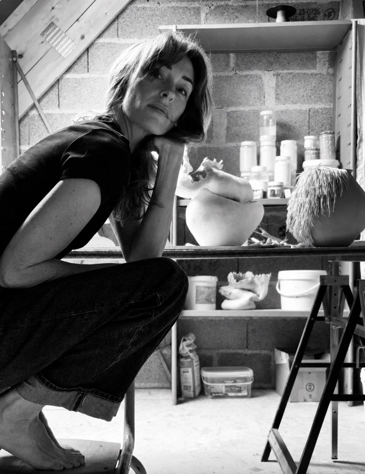 Une femme assise en train de sculpter des pichets en céramique dans un atelier, avec des matériaux et des outils visibles en arrière-plan.