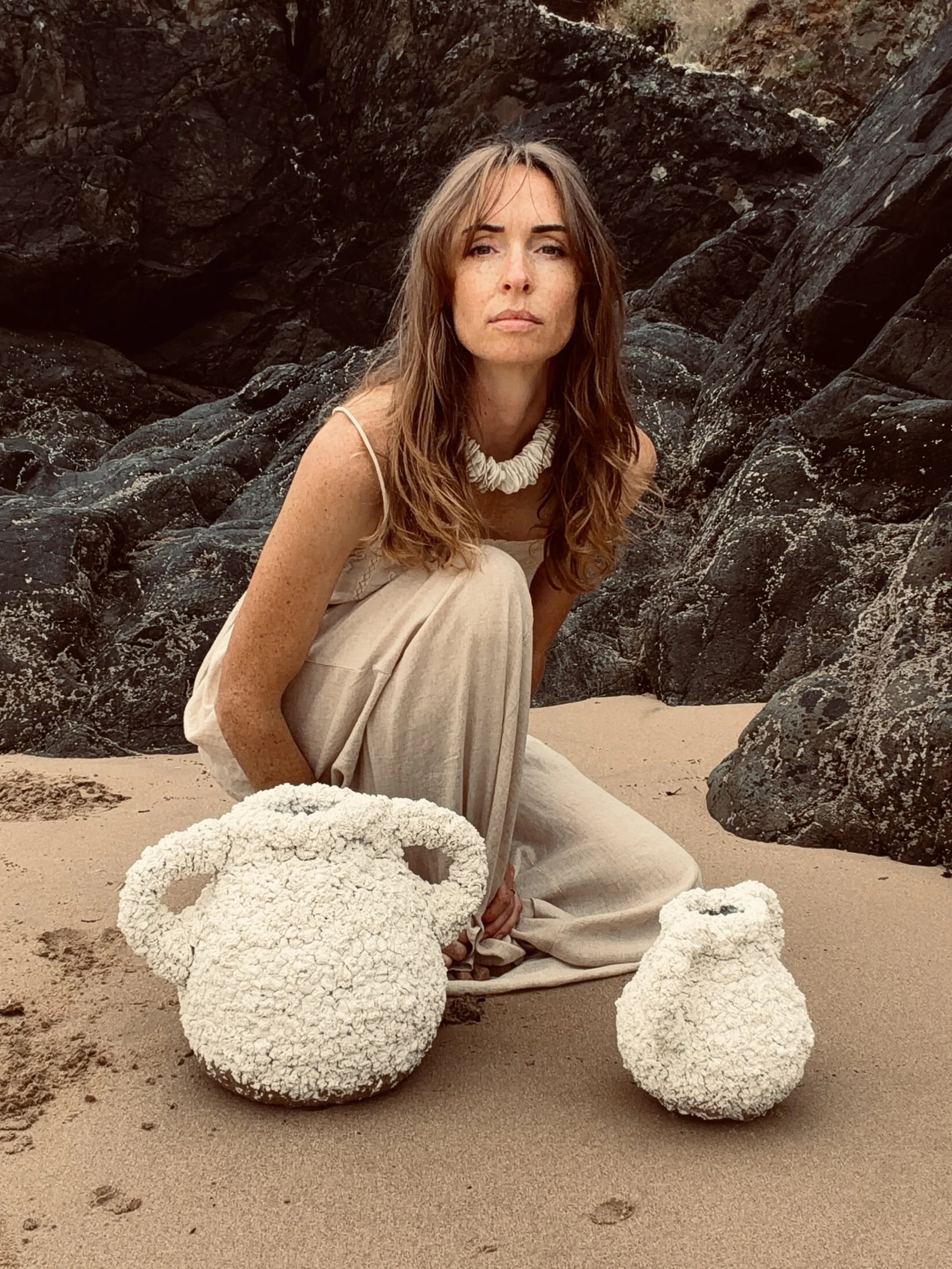 L'artiste est assise sur la plage entourée de rochers, portant une robe beige et un collier de perles en porcelaine, avec deux vase inspirées des coraux des mers froides.