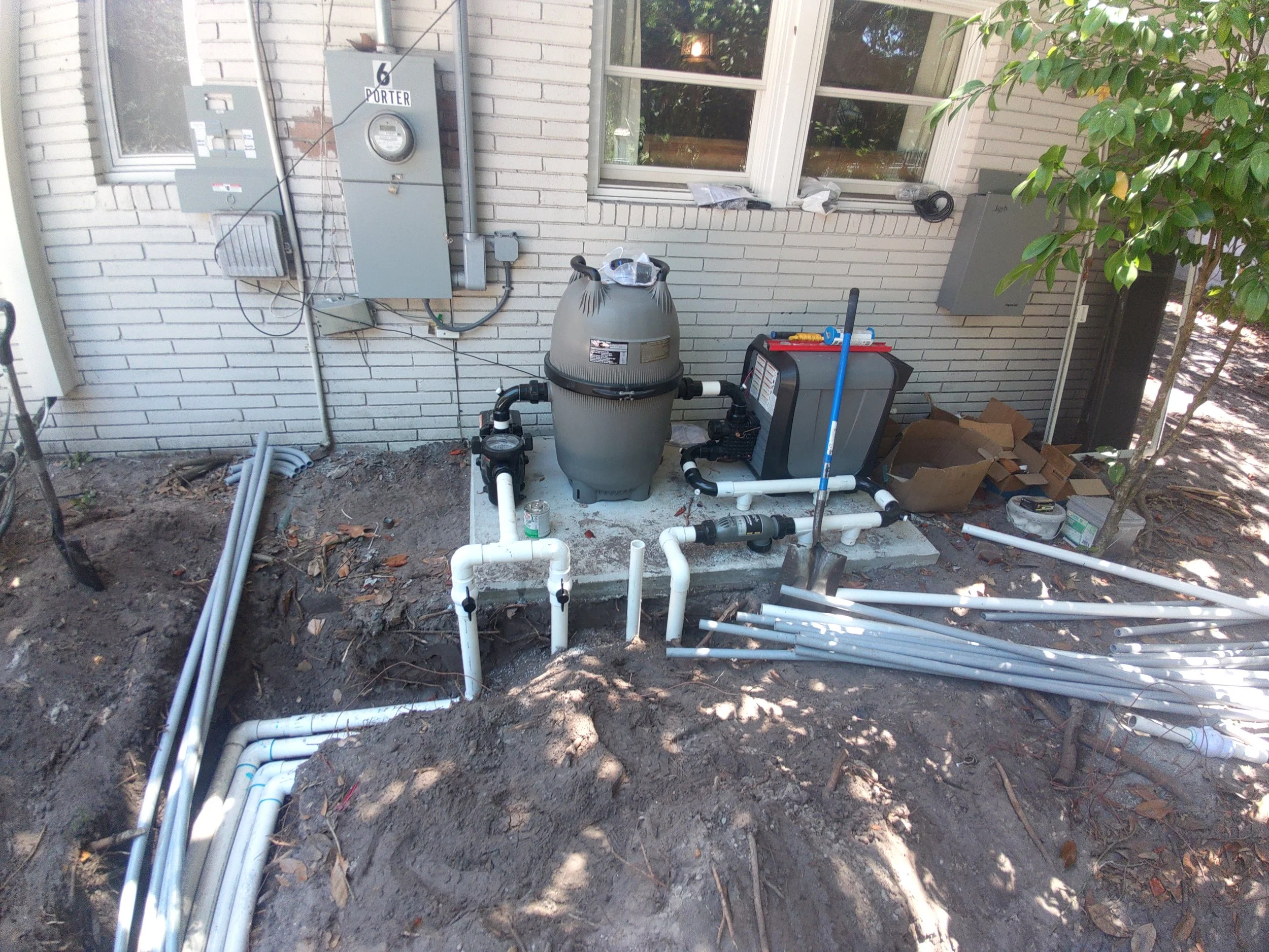 Installation of pool equipment outdoors, including a sand filter, pump, block of controls, and plumbing pipes, near a brick wall with electrical meters and gauges.