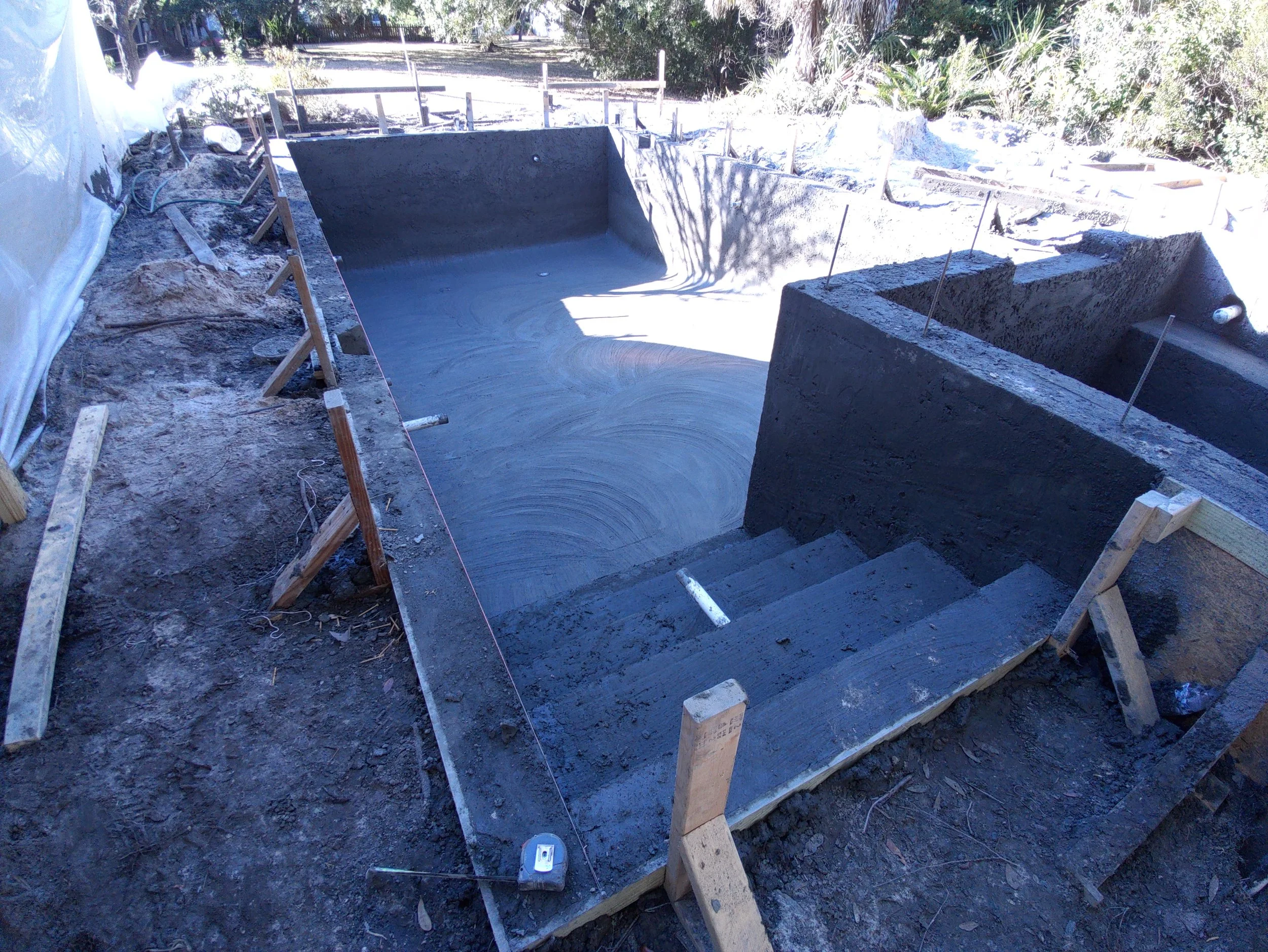 Construction site with a freshly poured concrete swimming pool and stairs, surrounded by wooden framing and construction tools.