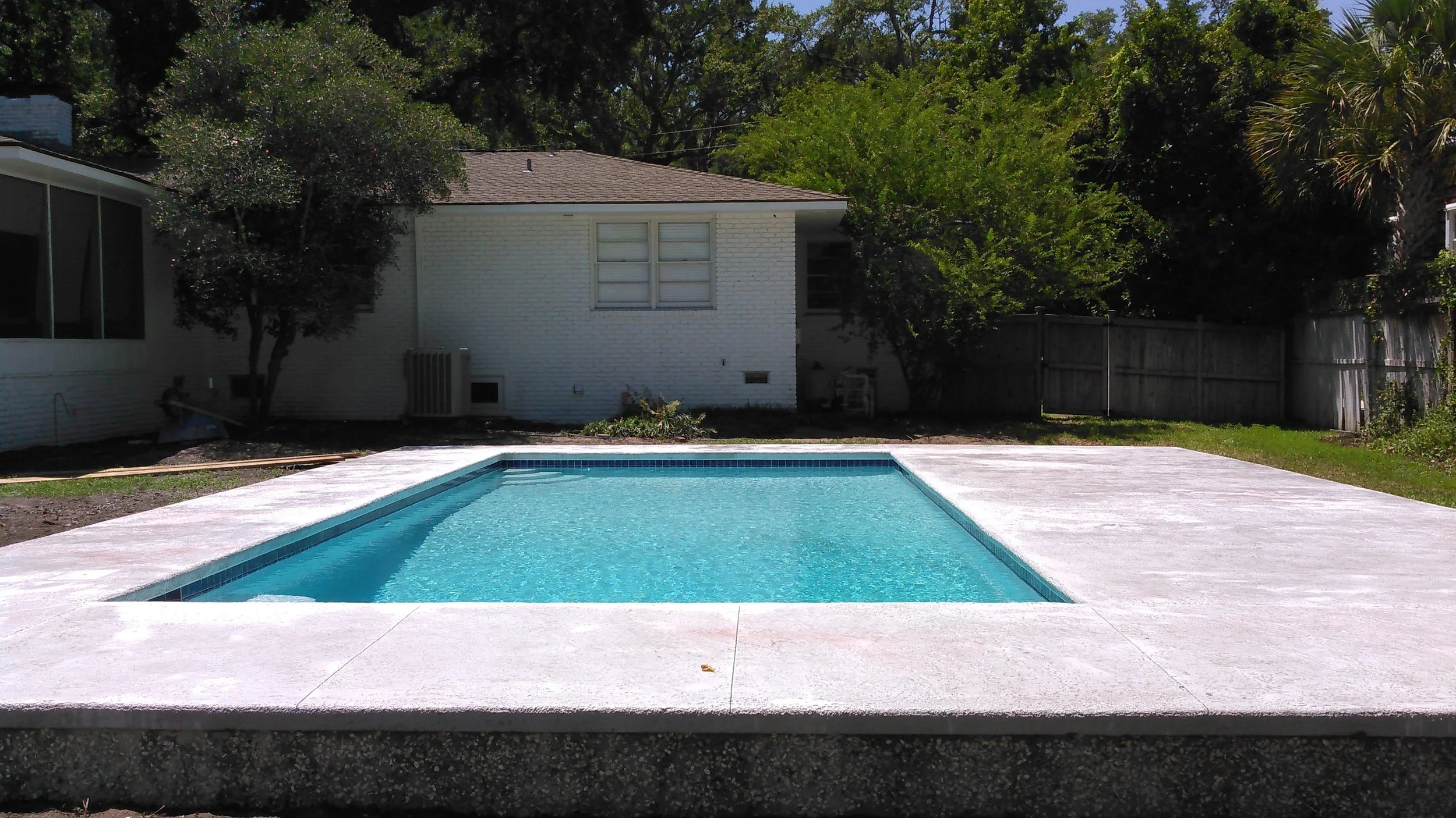 A backyard with a rectangular swimming pool surrounded by a white concrete deck, with a white house and trees in the background.