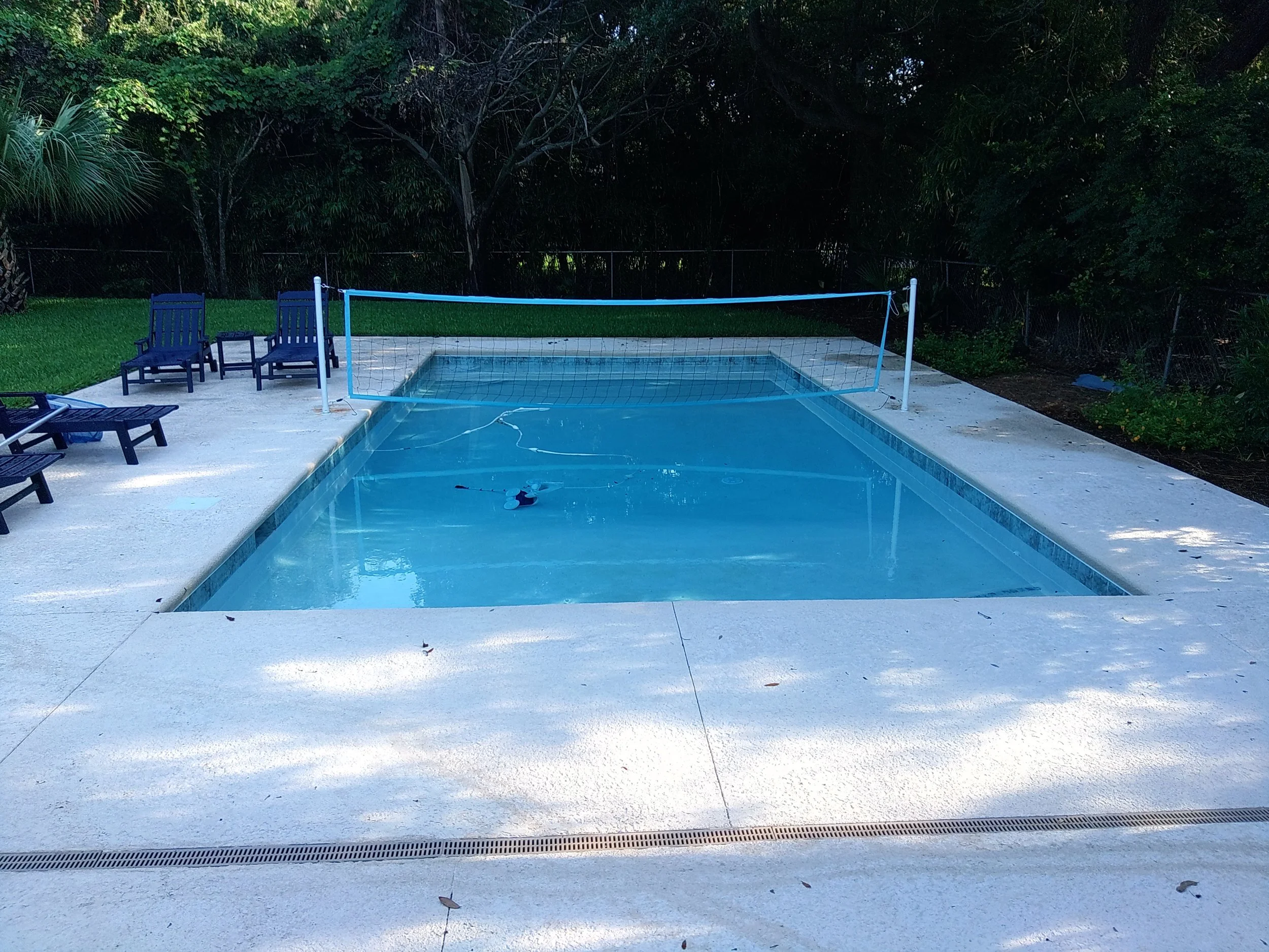 An empty swimming pool with clear blue water, surrounded by a white concrete deck with a small volleyball net set up at one end, and blue outdoor lounge chairs on the left side, with trees and bushes in the background.