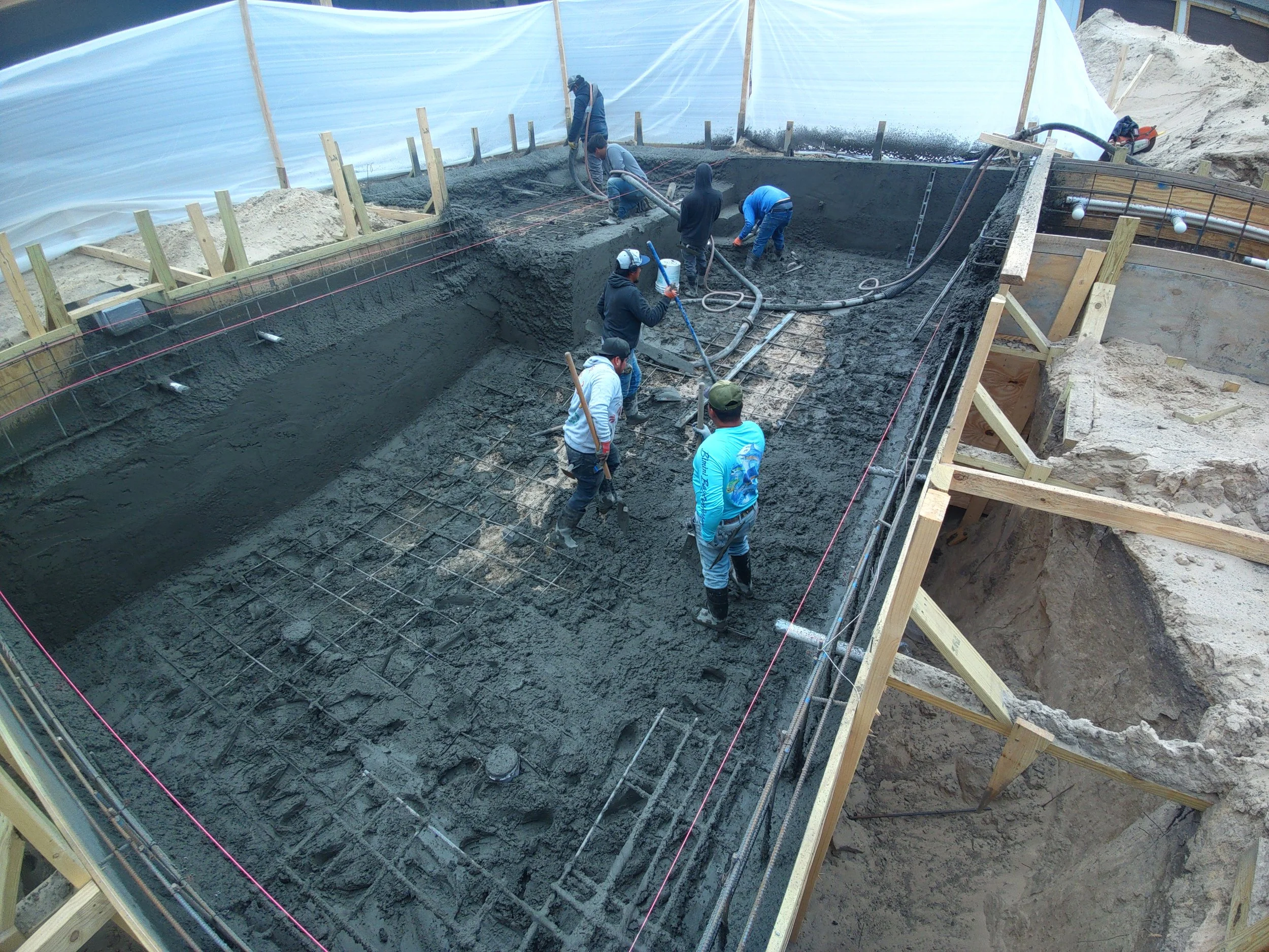 Construction workers pouring and spreading concrete in a building foundation, with wooden forms and rebar in place.