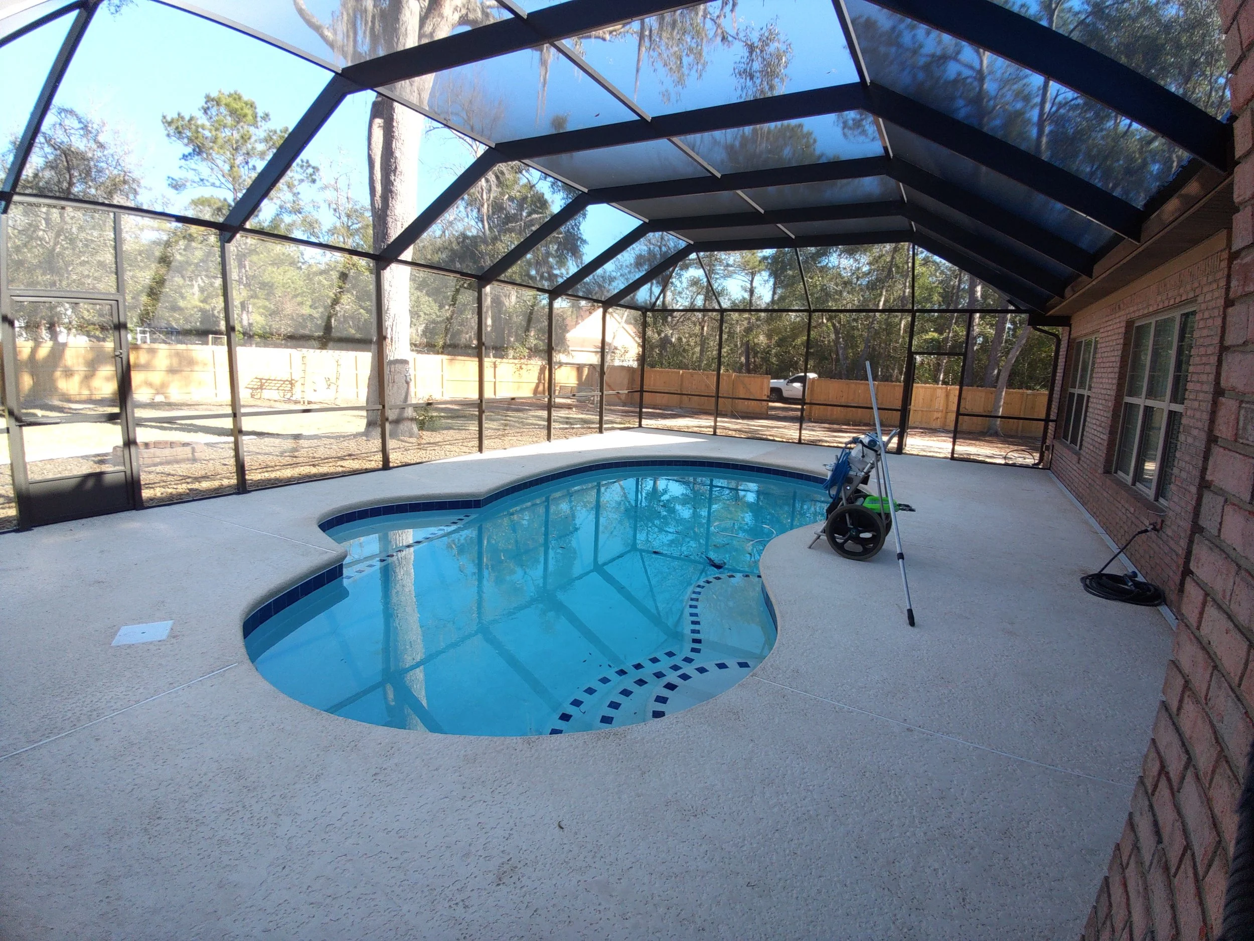 An enclosed backyard swimming pool area with a screened structure, a curved rectangular pool, a concrete deck, and a brick house wall on the right side.