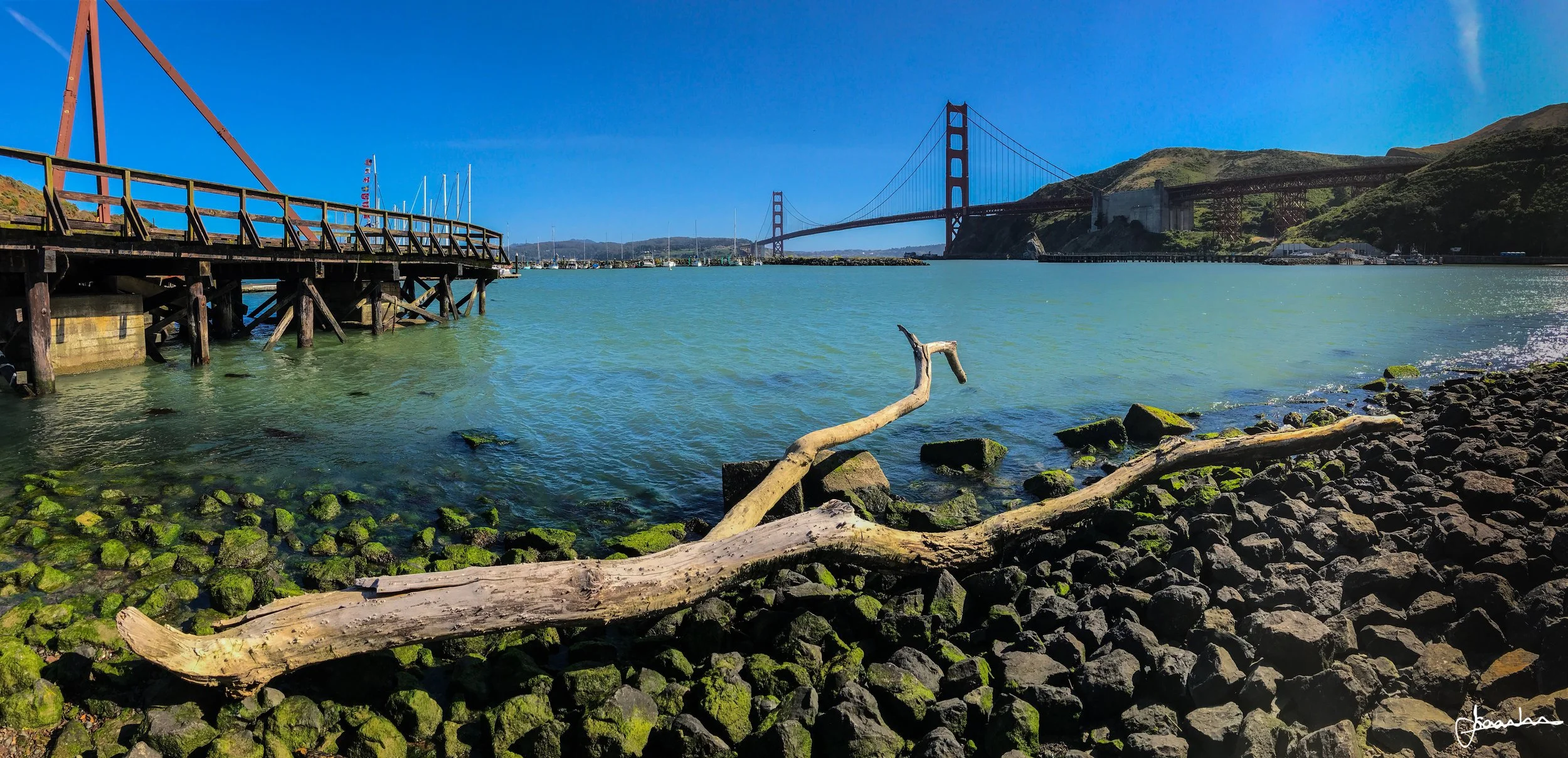 View of San Francisco Bay with the Golden Gate Bridge in the background, a wooden pier on the left, and rocky shoreline with driftwood in the foreground.