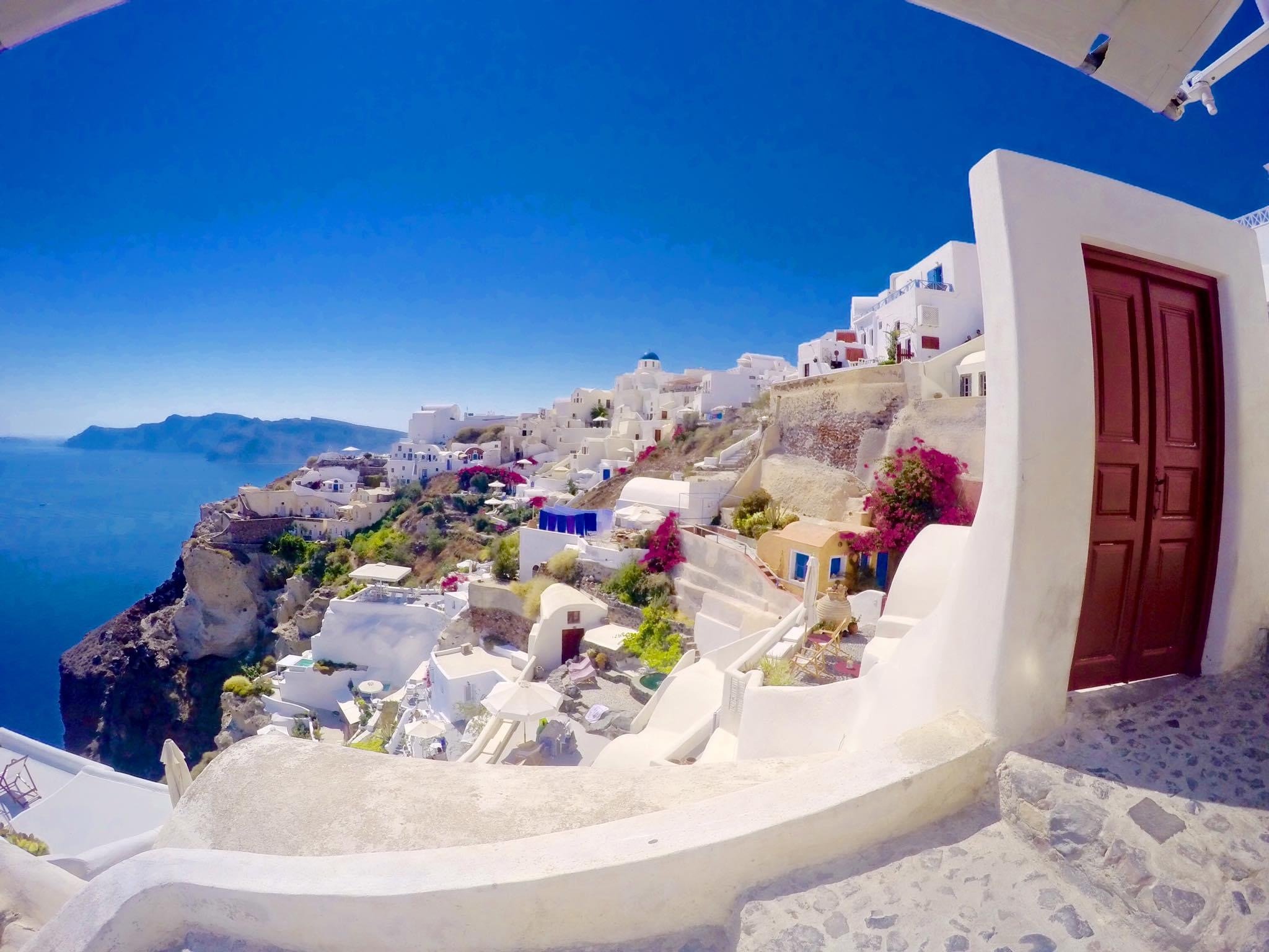 View of white buildings on a cliffside in Santorini, Greece, with blue sky and ocean in the background, and a wooden door in the foreground.