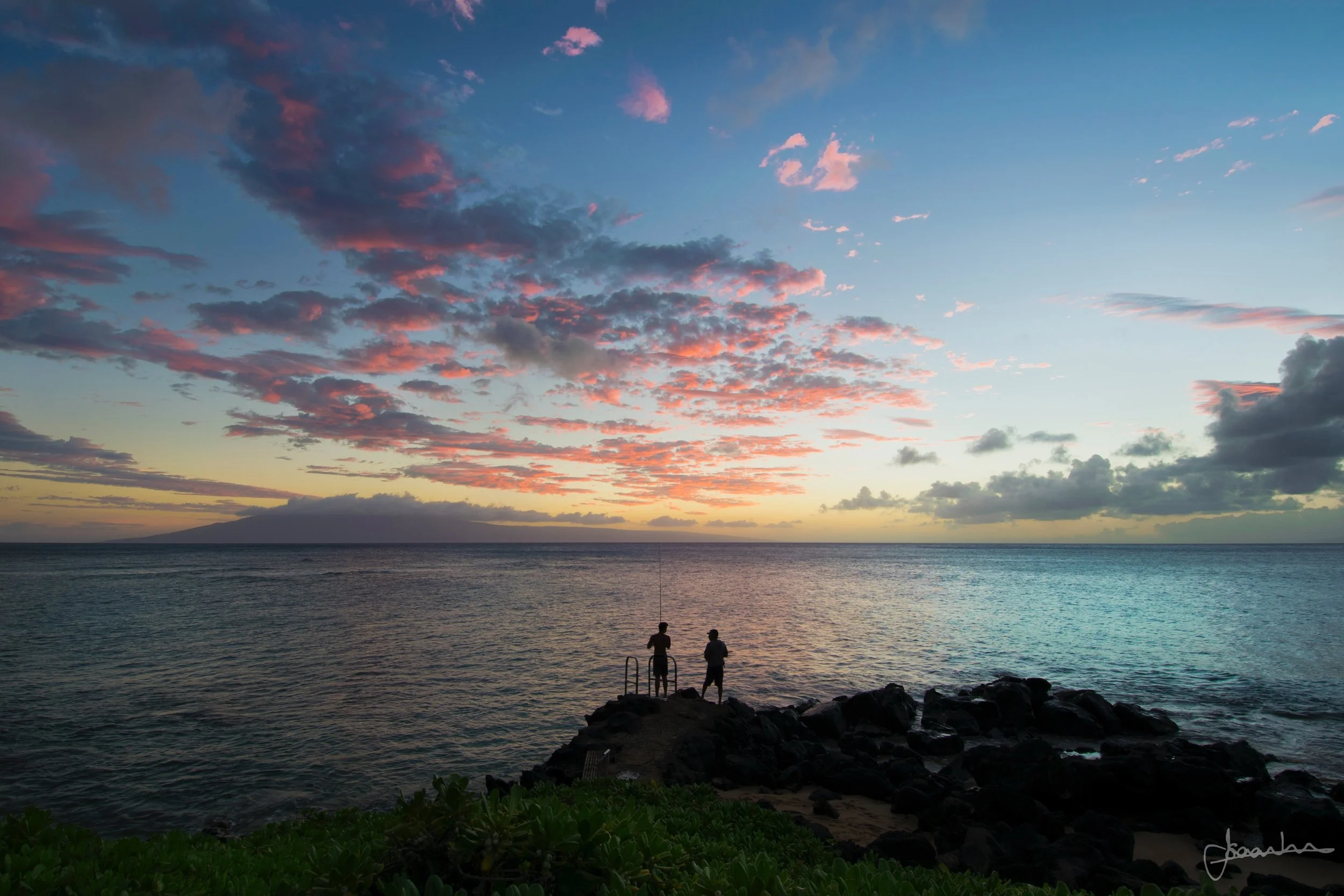 A sunset over the ocean with scattered clouds and two people standing on rocks fishing, silhouetted against the colorful sky.