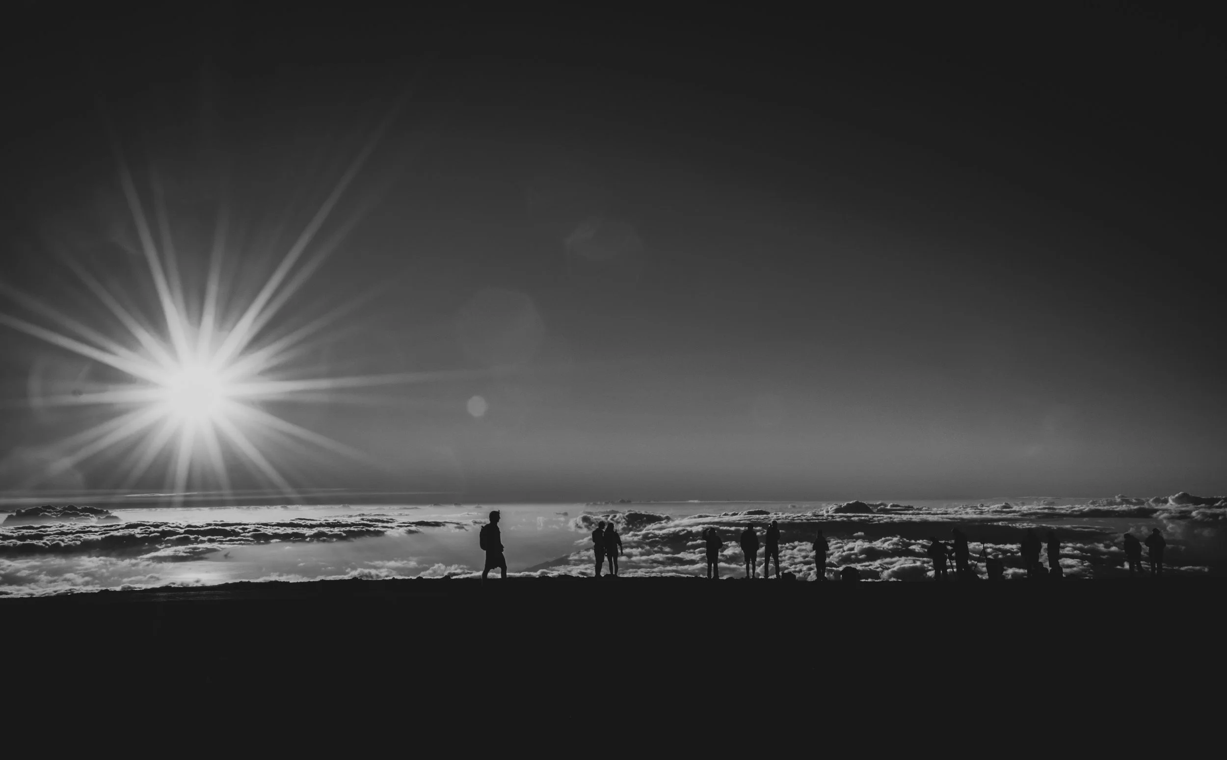 Silhouettes of people standing on a hilltop under a bright sun with rays, clouds in the background, in black and white.