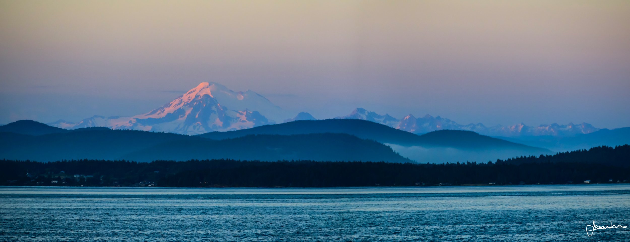 Scenic view of mountains with snow-capped peaks in the distance, layered with forested hills in the foreground, over a body of water at dusk or dawn.