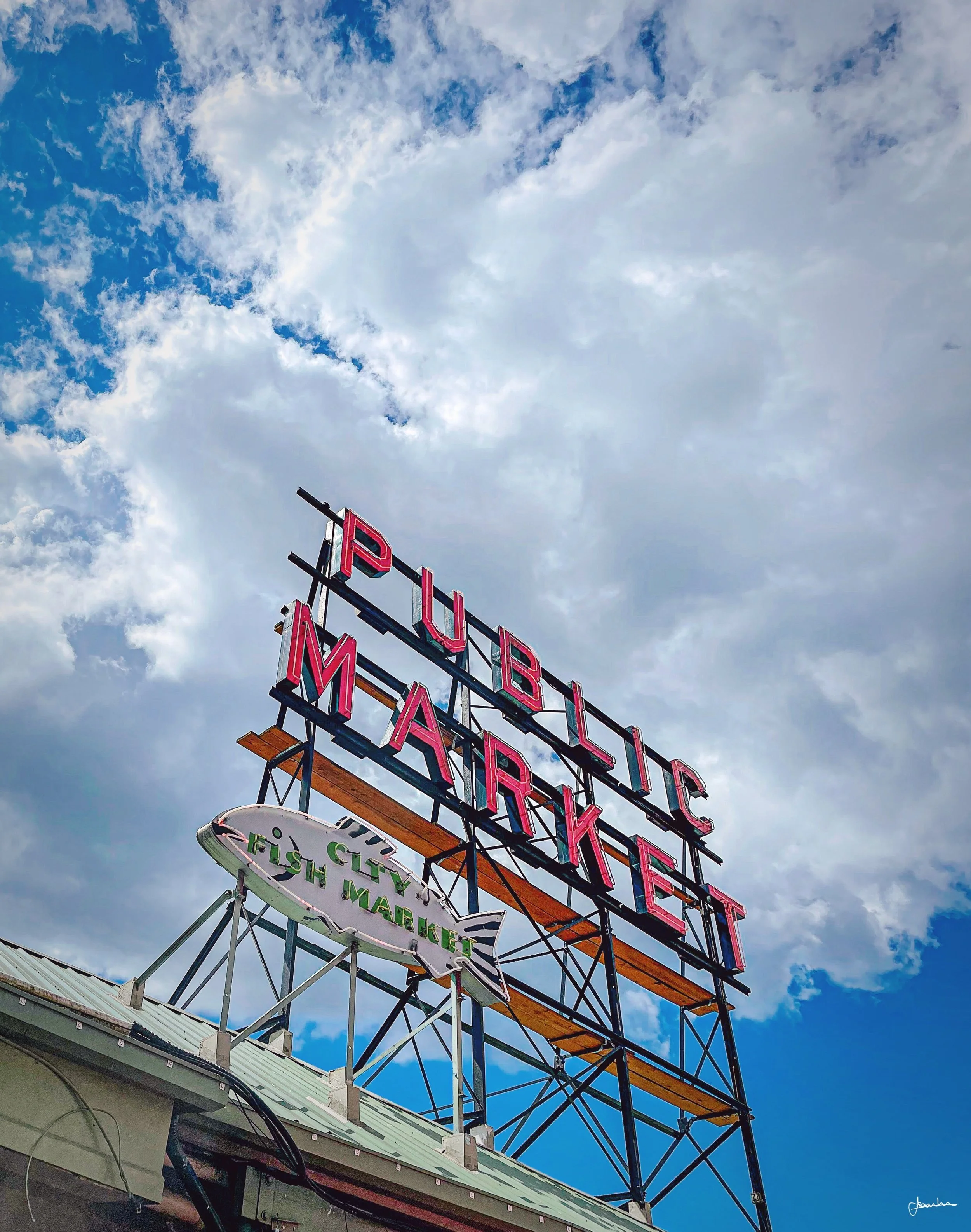 Bright pink neon sign reads 'Public Market' above a smaller sign shaped like a fish that says 'Fish Market', set against a partly cloudy sky.