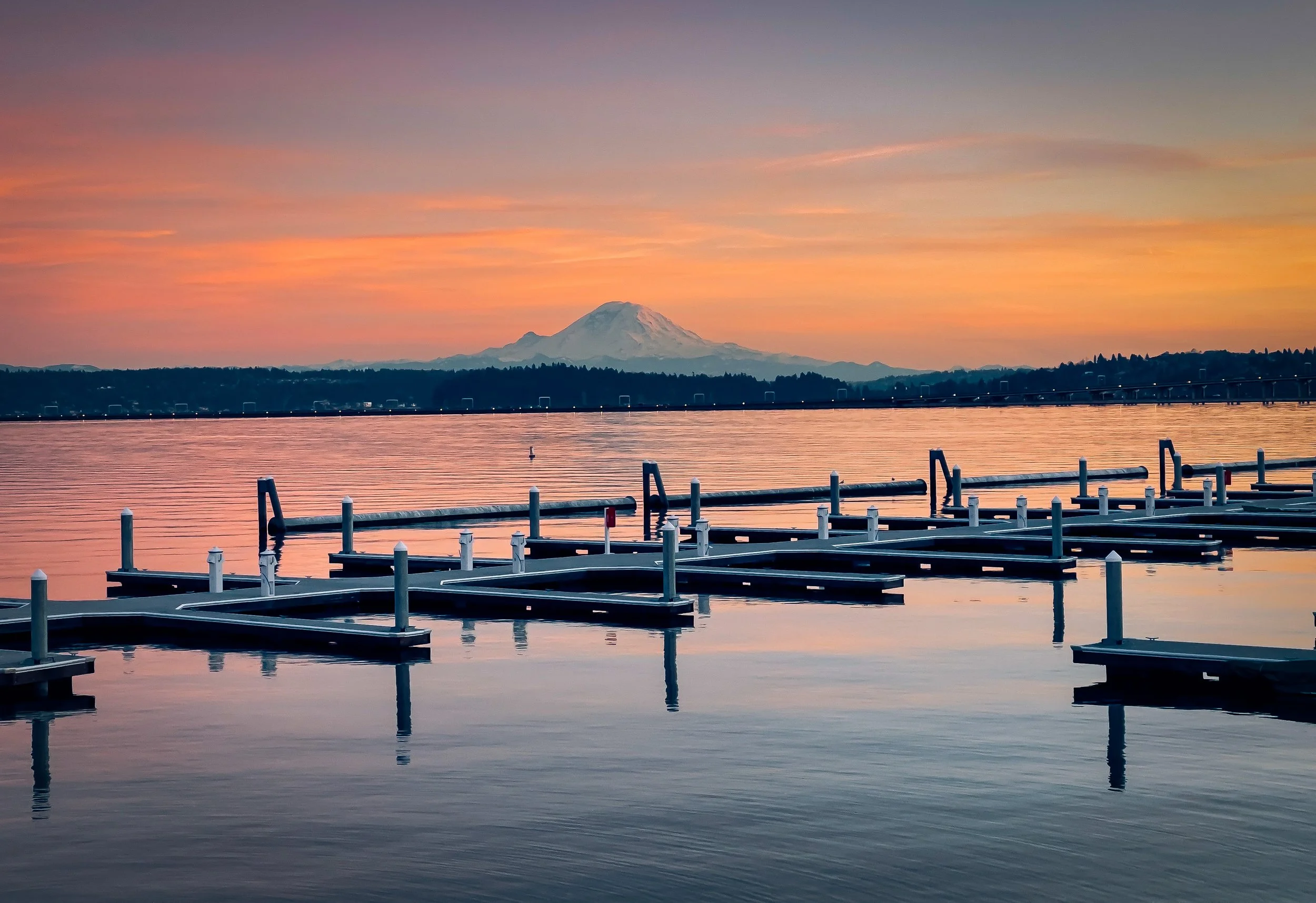 A lake with empty docks extends into the water at sunset, with Mount Fuji visible in the background and a colorful sky overhead.
