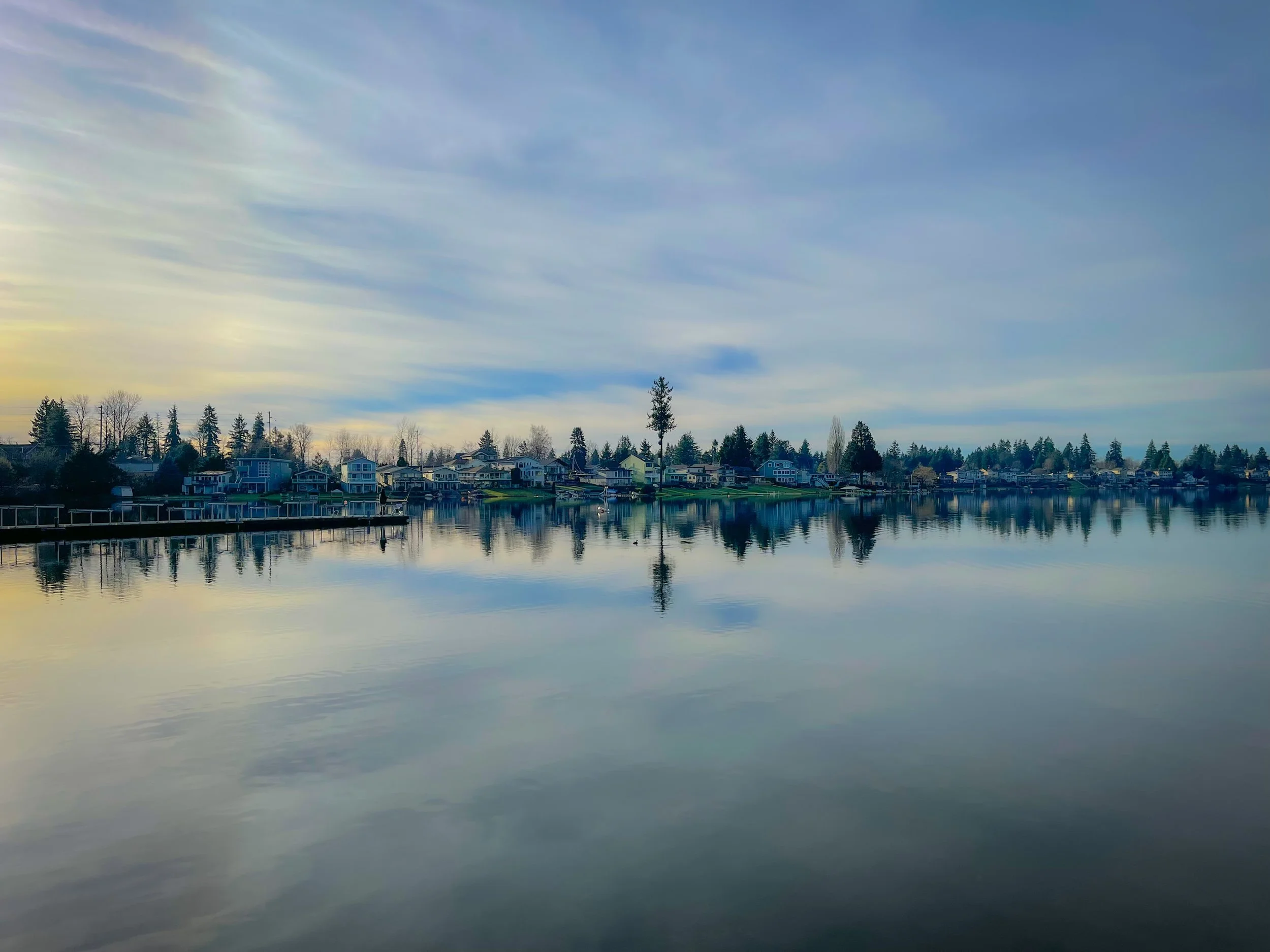 A peaceful lake scene with houses along the shoreline, calm water reflecting the sky and houses, with a partly cloudy sky and a distant treeline.