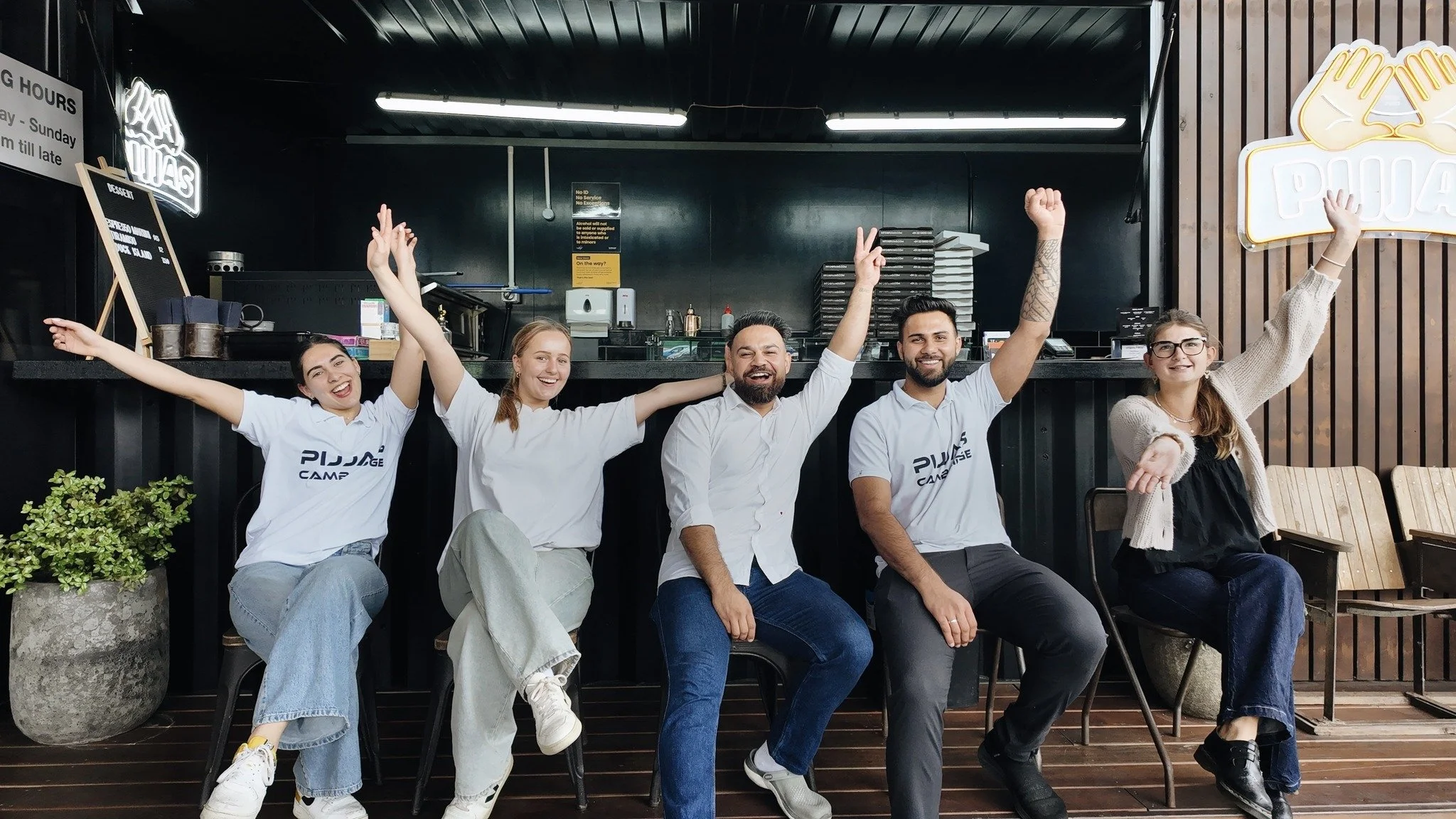 Five happy people sitting and standing in a coffee shop, raising their hands and smiling. Four of them are wearing white t-shirts with the text 'Pijjae Camp'.