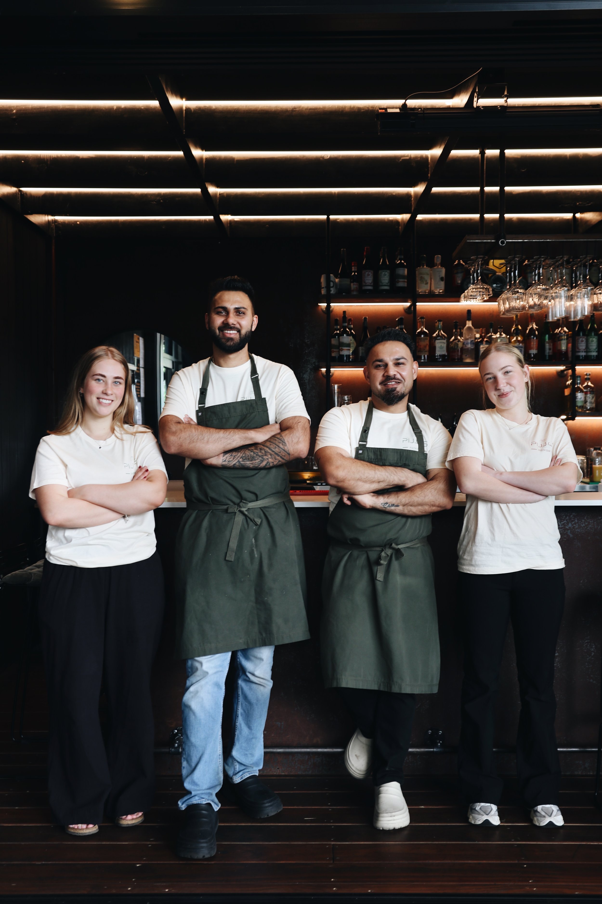 Group of four restaurant staff members standing with arms crossed in front of bar, smiling, with bottles and glasses behind them.