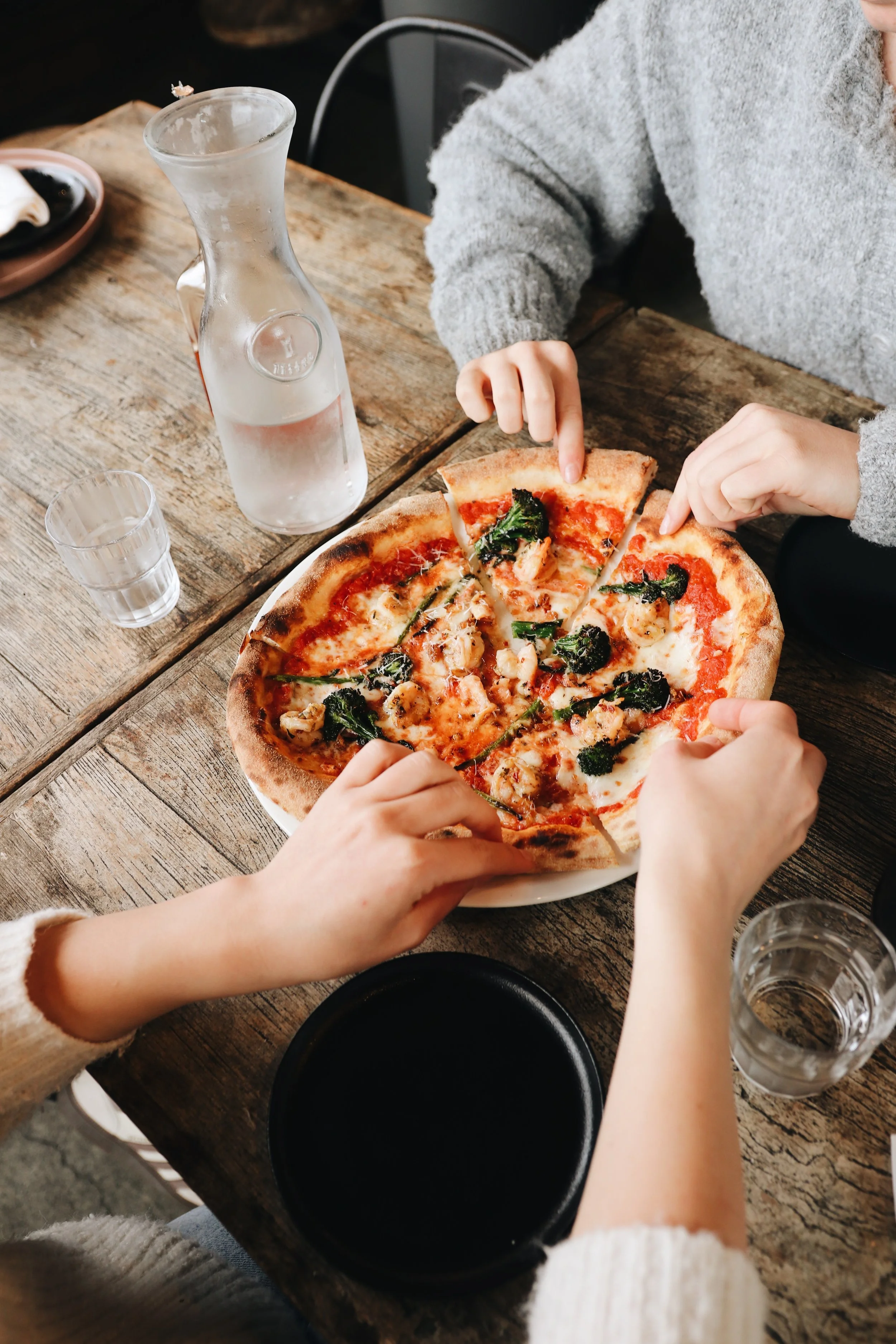 A person in a gray sweater is slicing a pizza topped with meats, greens, and cheese on a rustic wooden table. There is a glass water bottle, an empty glass, and a black plate nearby.