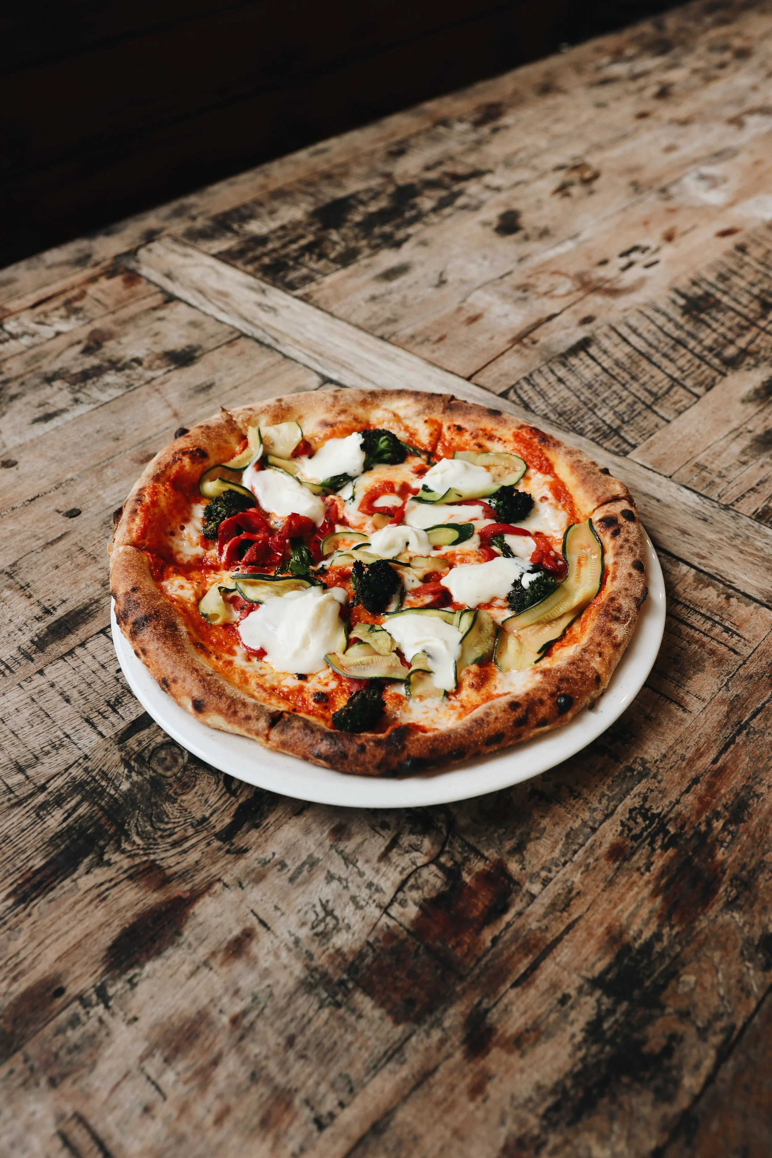 A pizza with vegetables on a white plate, placed on a rustic wooden table.