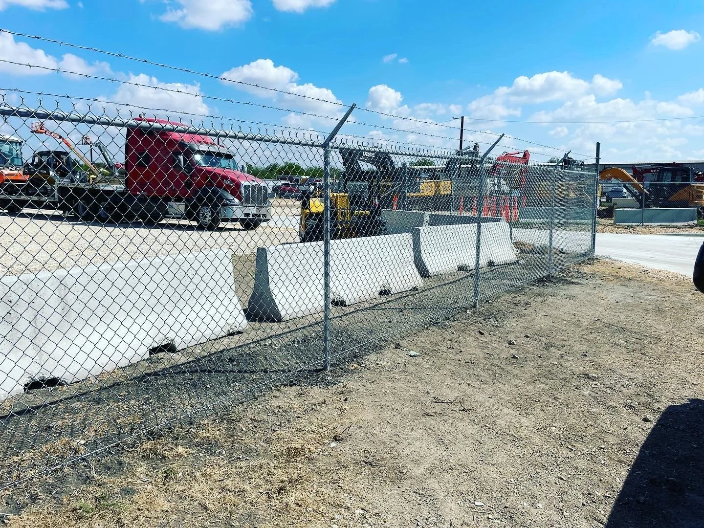 Commercial site with heavy machinery like bulldozers and trucks, surrounded by a chain-link fence that we built, under a partly cloudy sky.